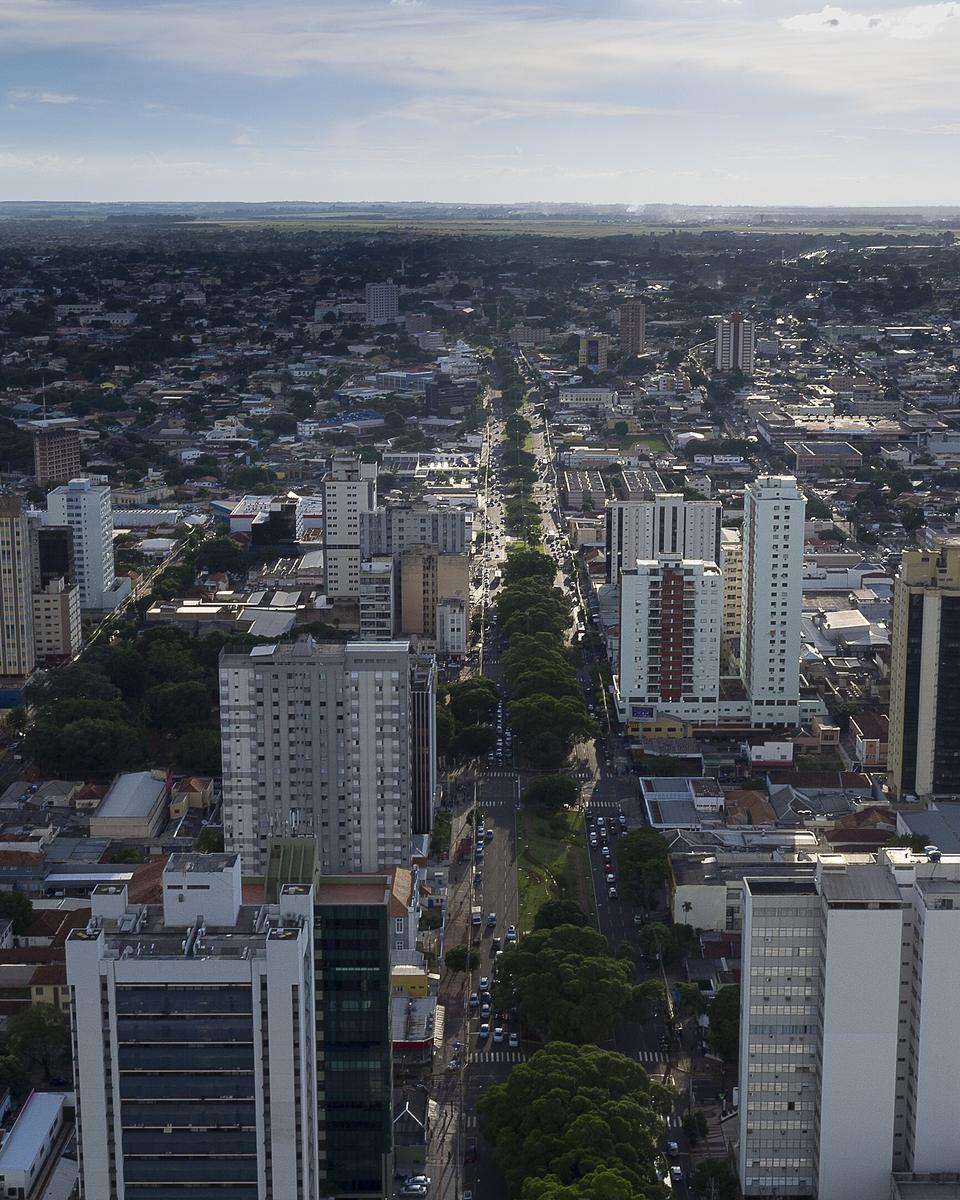 Vista aérea da Avenida Mato Grosso cortando Campo Grande em linha reta até o horizonte, com fileira de árvores no meio da pista e prédios altos e baixos misturados nos dois lados.