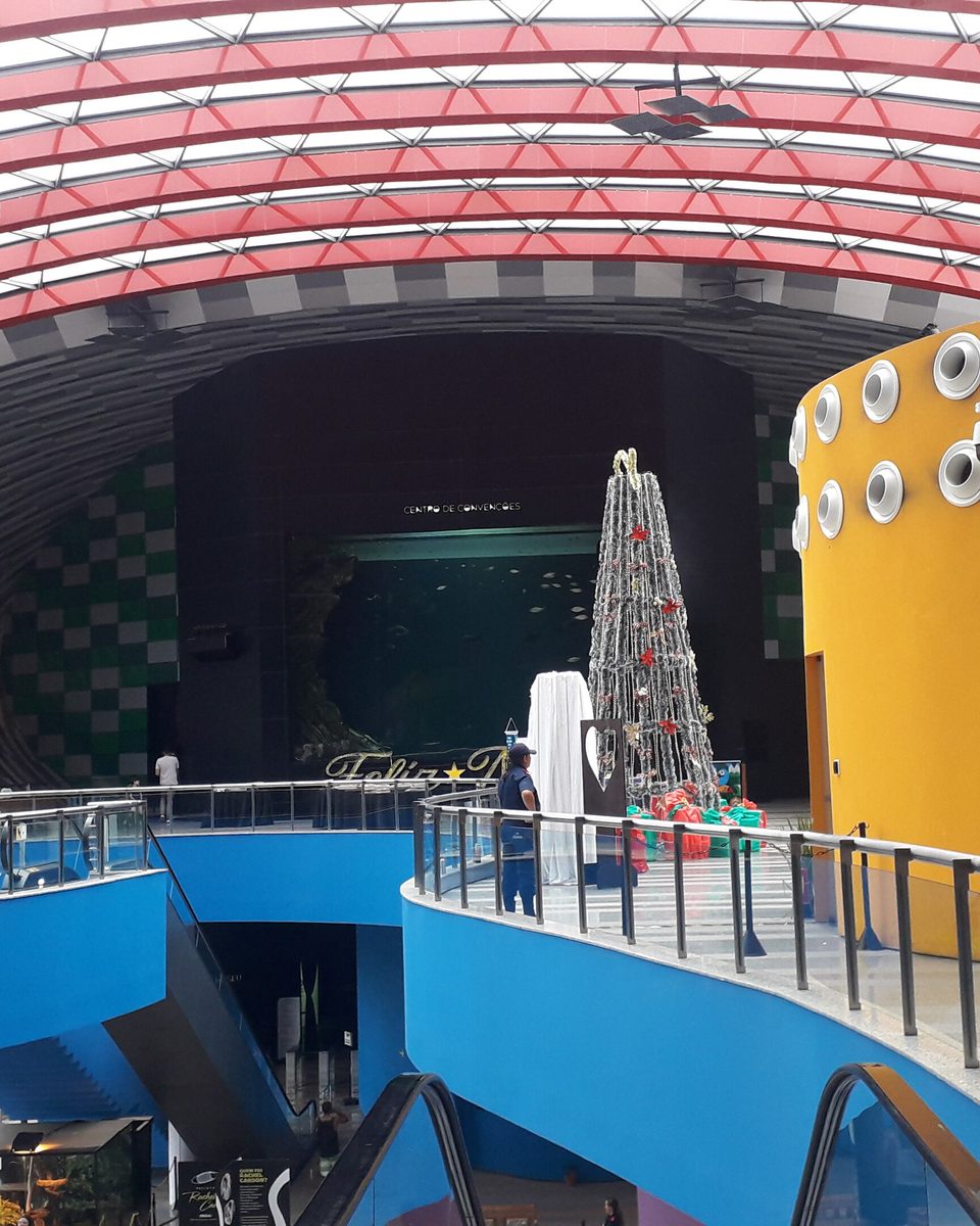 Fachada curva do Bioparque Pantanal, com a cobertura listrada em vermelho e branco, o volume amarelo com janelas redondas e visitantes nas passarelas azuis.