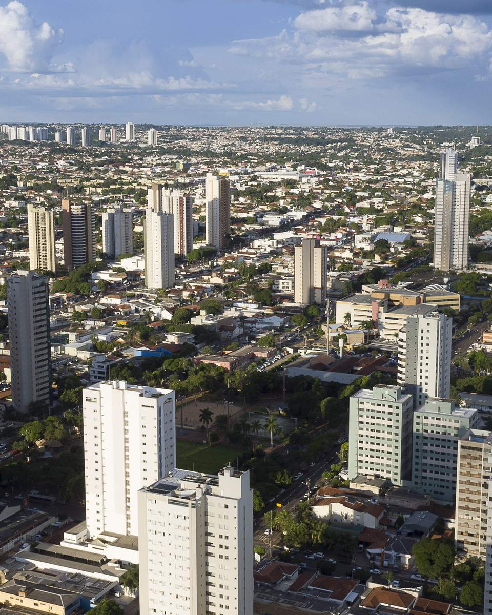 Vista aérea de Campo Grande com prédios altos em primeiro plano, a cidade se estendendo plana até o horizonte e nuvens brancas cobrindo o céu azul.