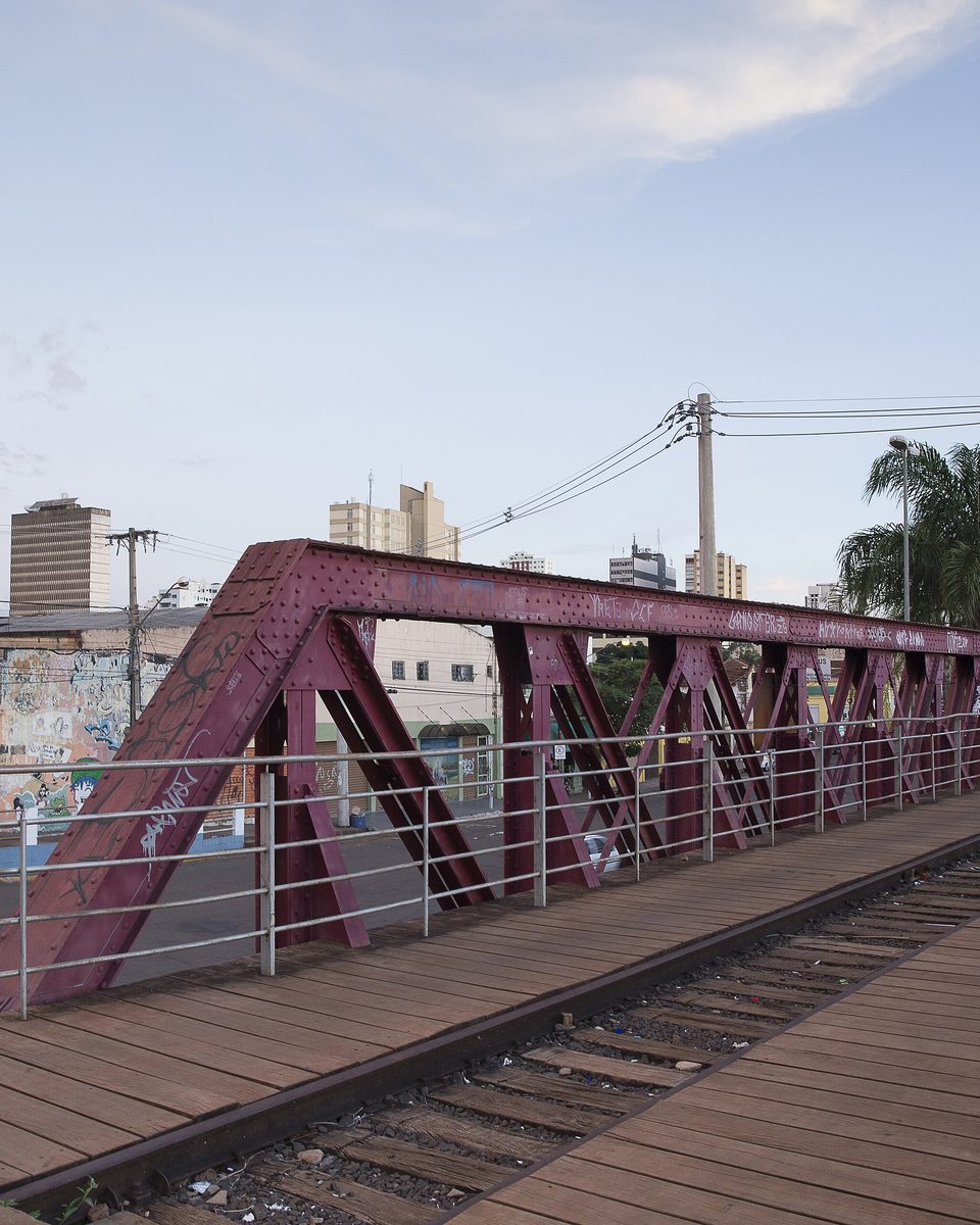 Viaduto de estrutura metálica vermelha sobre a linha férrea em Campo Grande, com muro grafitado, prédios do centro ao fundo e céu de fim de tarde.
