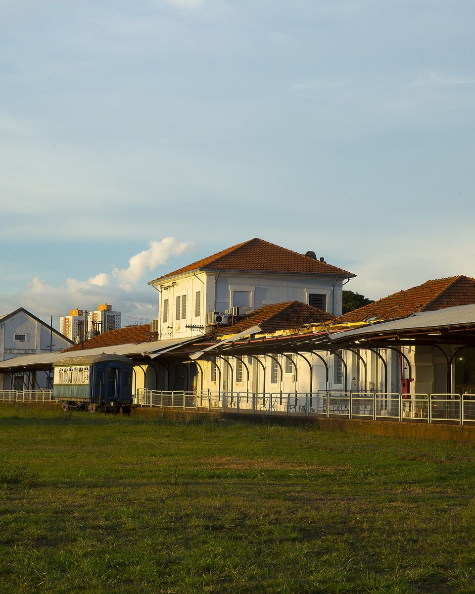 Galpões e plataforma da antiga Estação Esplanada, em Campo Grande, iluminados pelo sol do fim da tarde, com um vagão azul nos trilhos e prédios da cidade ao fundo.