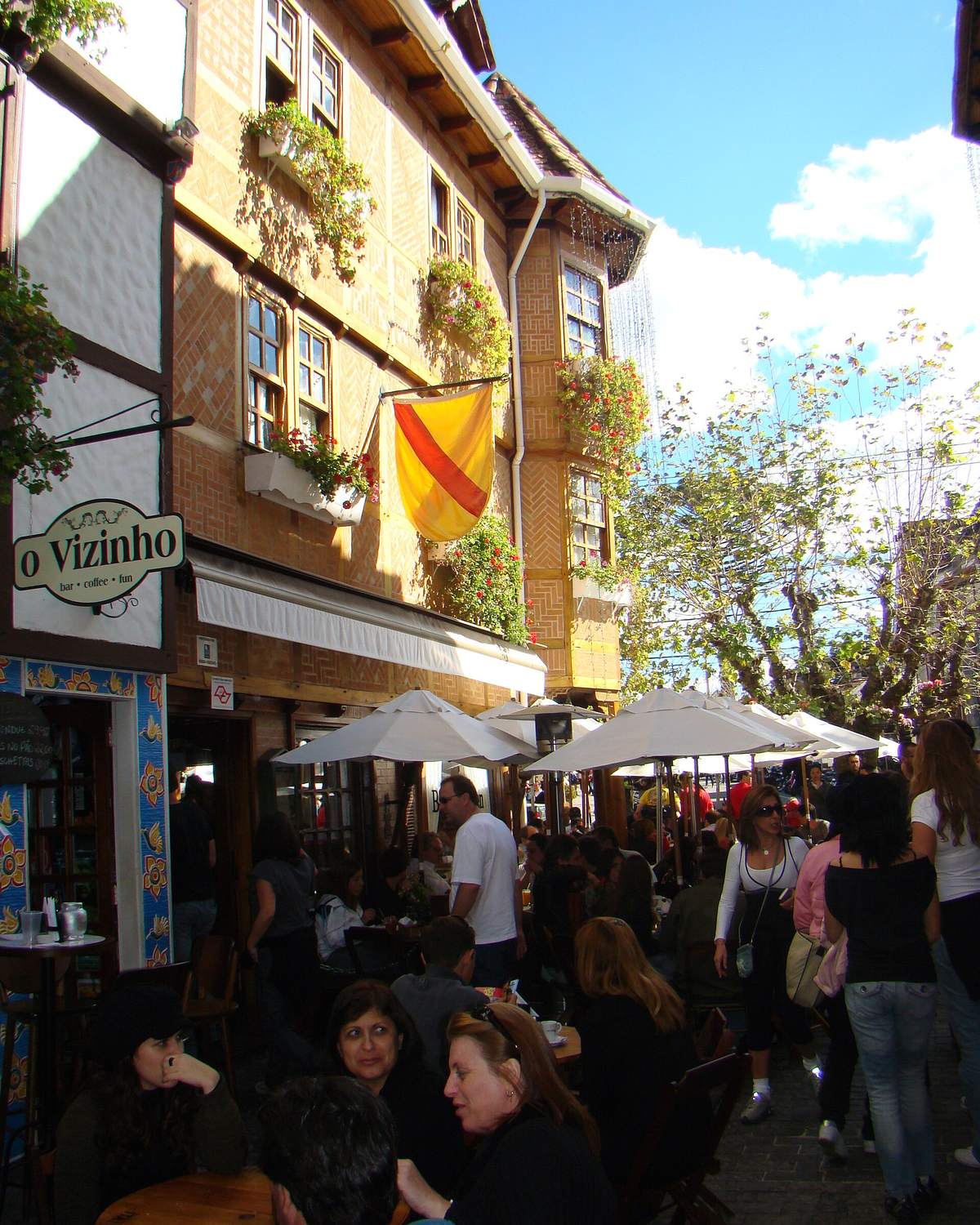 Rua de pedestres da Vila Capivari cheia de turistas agasalhados, com mesas de restaurante, guarda-sóis brancos e casas de estilo europeu com flores nas janelas.