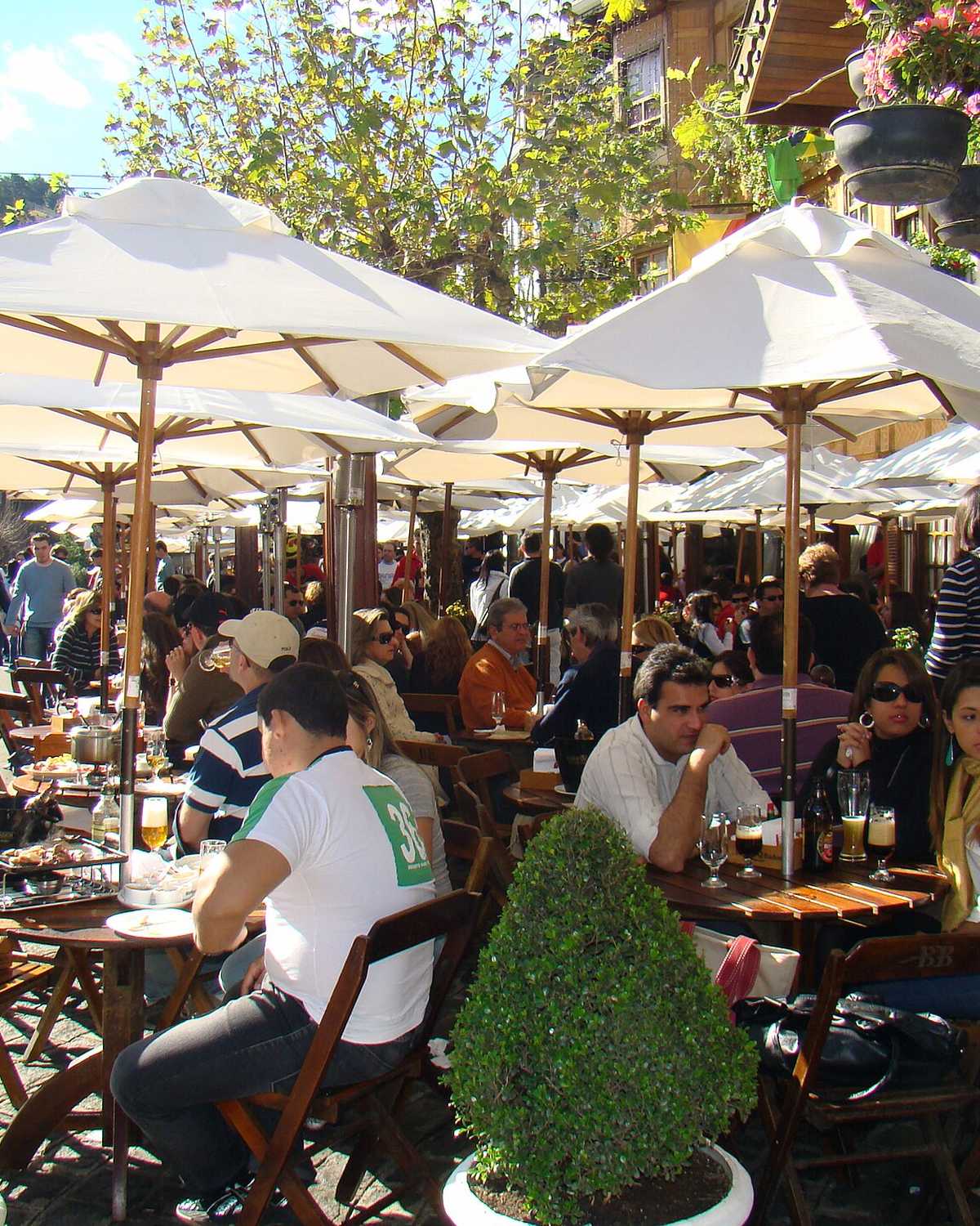 Mesas ao ar livre lotadas sob guarda-sóis brancos numa praça de Campos do Jordão, com casais e amigos almoçando entre vasos de plantas.