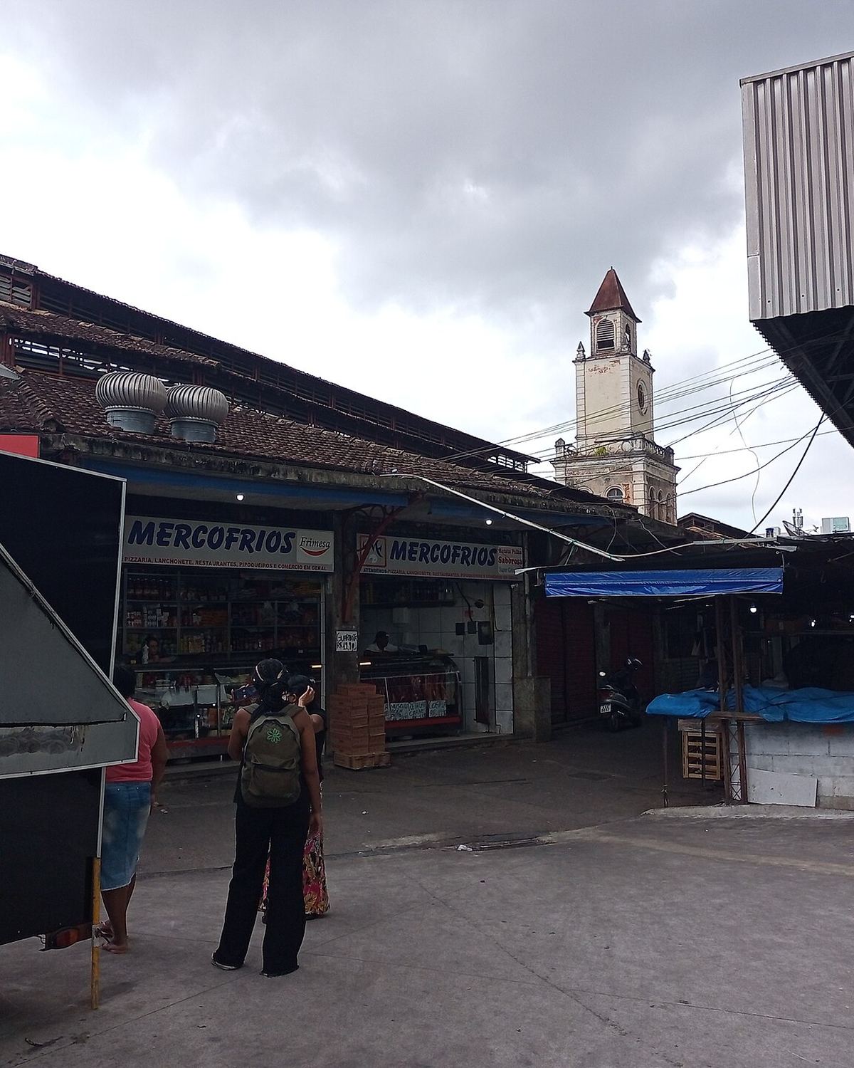 Mulheres em frente às lojas do Mercado Municipal de Campos dos Goytacazes, com a torre de uma igreja ao fundo.