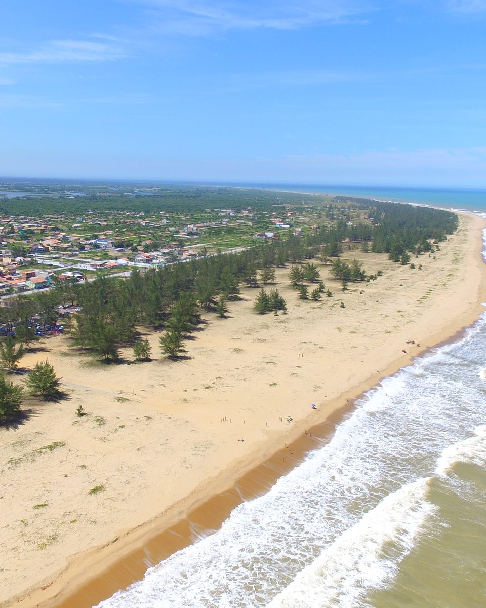 Vista aérea da praia de Farol de São Thomé, em Campos dos Goytacazes, com a faixa de areia clara fazendo uma curva, ondas quebrando à direita e o bairro com casas e coqueiros à esquerda.