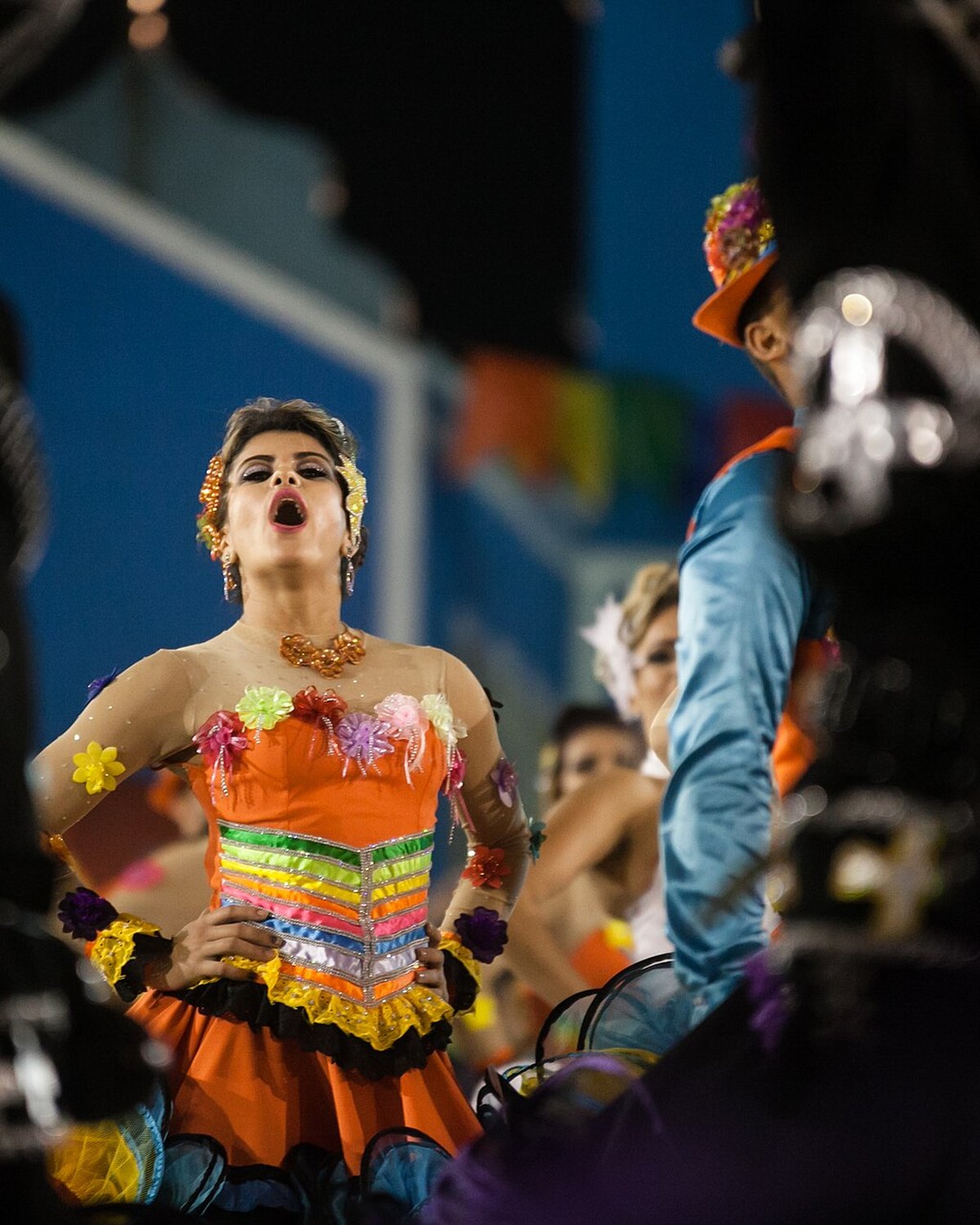 Dançarina de quadrilha junina de vestido laranja e colorido com flores, cantando de mãos na cintura entre brincantes vestidos de preto, à noite no São João de Caruaru.