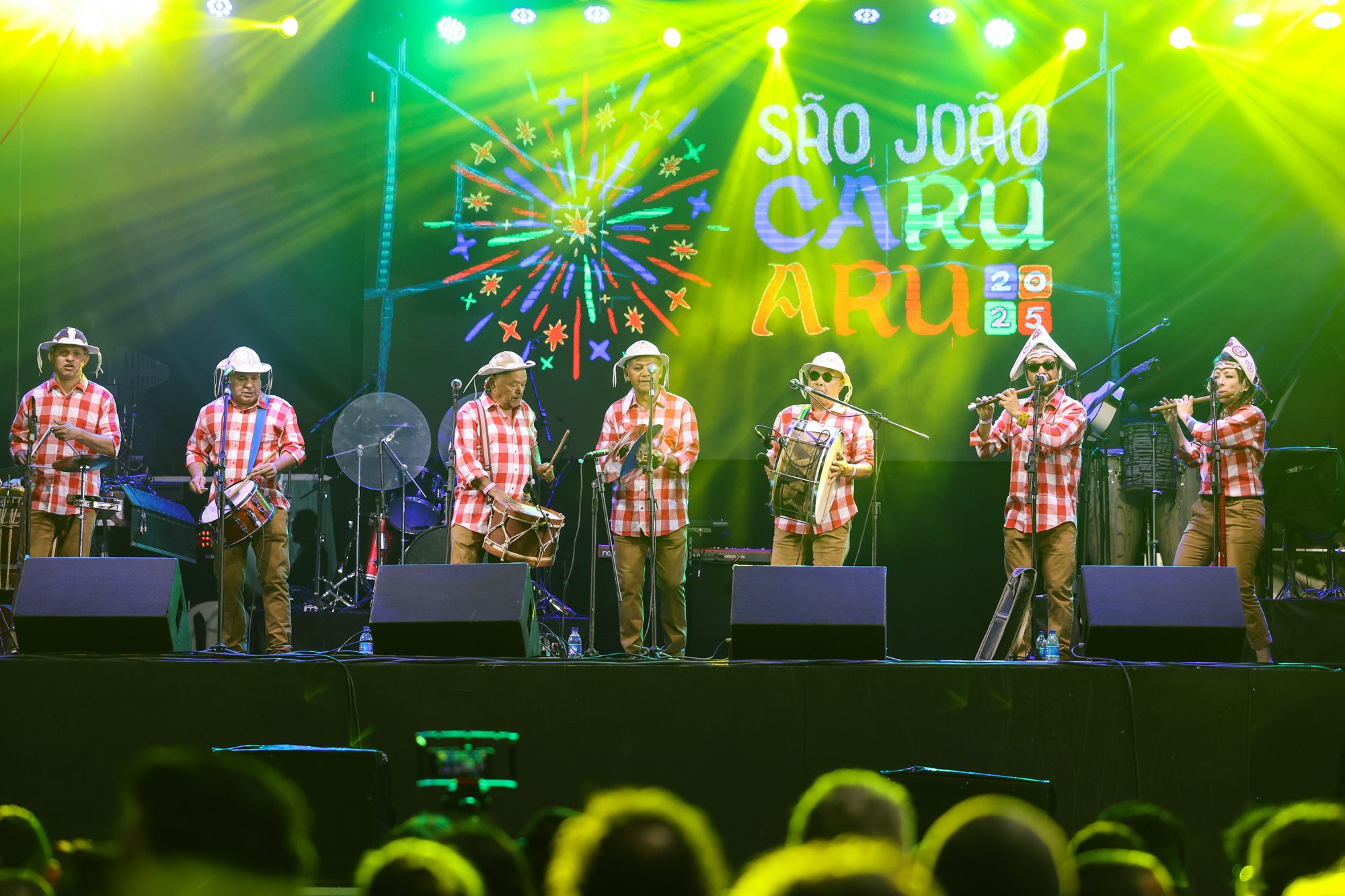 Banda de Pífanos de Caruaru tocando num palco do São João, com os músicos de camisa xadrez vermelha e chapéu de palha enfileirados, luzes verdes e amarelas e o painel São João Caruaru 2025 ao fundo, diante da plateia.