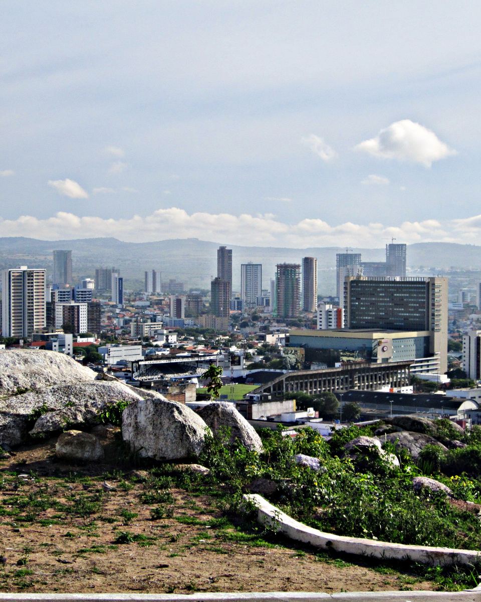 Vista de Caruaru a partir do Monte do Bom Jesus, com pedras e mato em primeiro plano, a cidade de casas baixas e os prédios do Centro até a serra no horizonte, sob céu azul com nuvens.