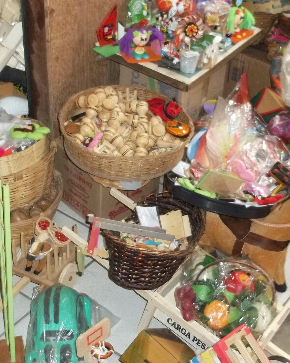 Banca de artesanato na Feira de Caruaru com cestos de palha, brinquedos de madeira, violinhas coloridas, bonecos e peças de barro amontoados no chão e nas prateleiras.