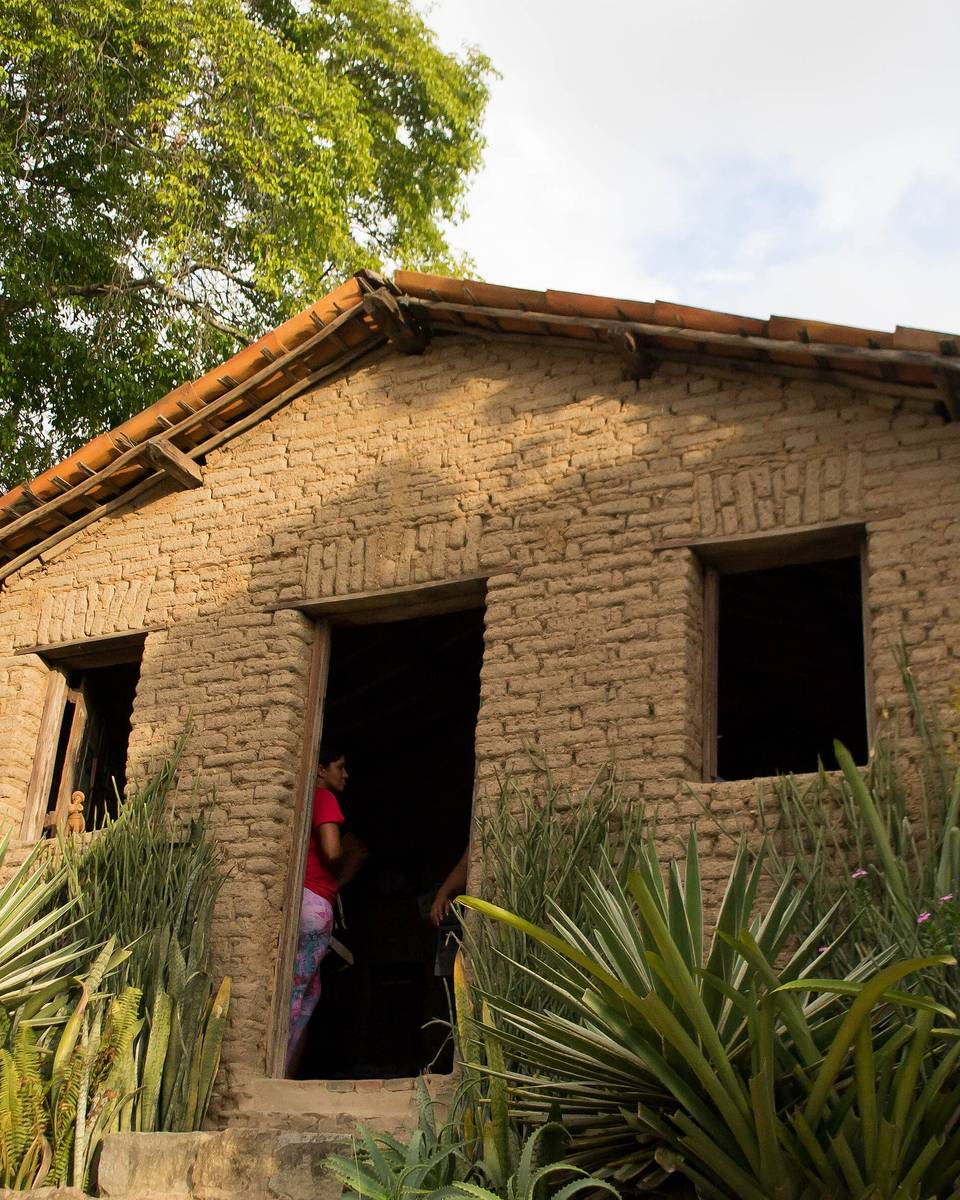Casa de taipa de Mestre Vitalino no Alto do Moura, de paredes de barro e telhado de telha, cercada de agaves e árvores, com um visitante de costas olhando a fachada.