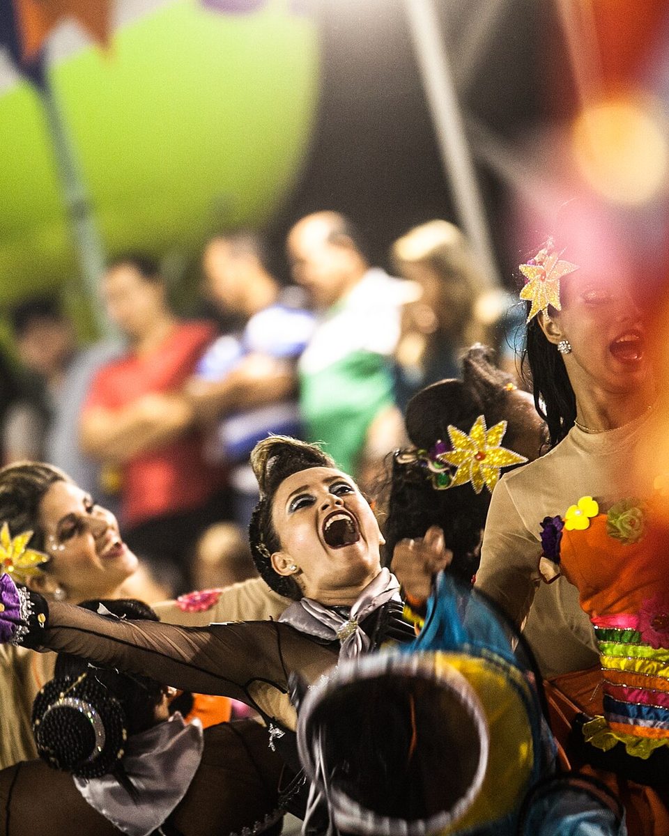 Quadrilha junina se apresentando à noite no São João de Caruaru, com dançarinas de vestidos coloridos e flores no cabelo em primeiro plano e o público atrás, sob luzes e bandeirinhas.