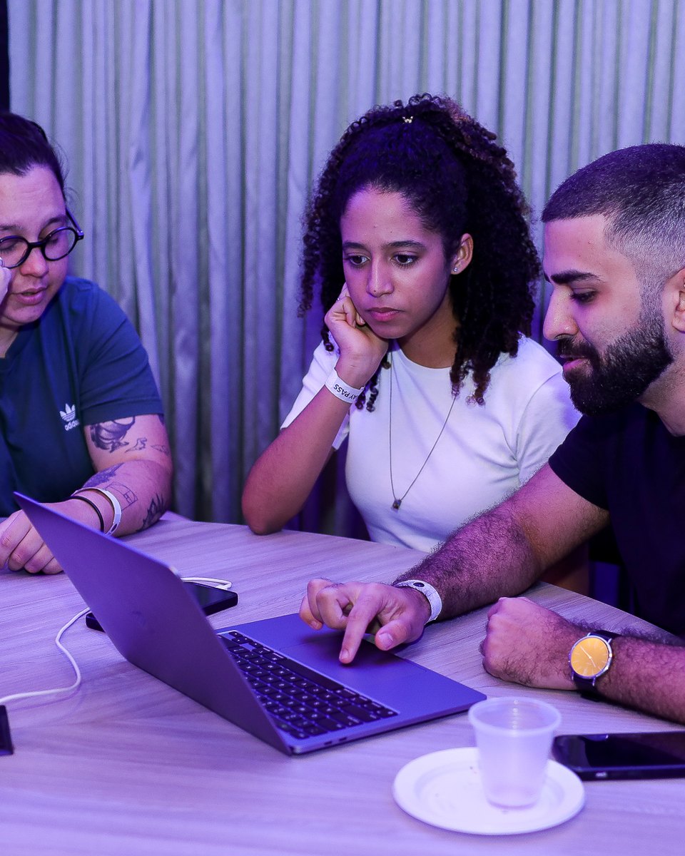 Três jovens em volta de um notebook numa mesa redonda durante a Campus Party Day Caruaru, uma de óculos com a mão no rosto, uma de camiseta branca e um rapaz de barba digitando.