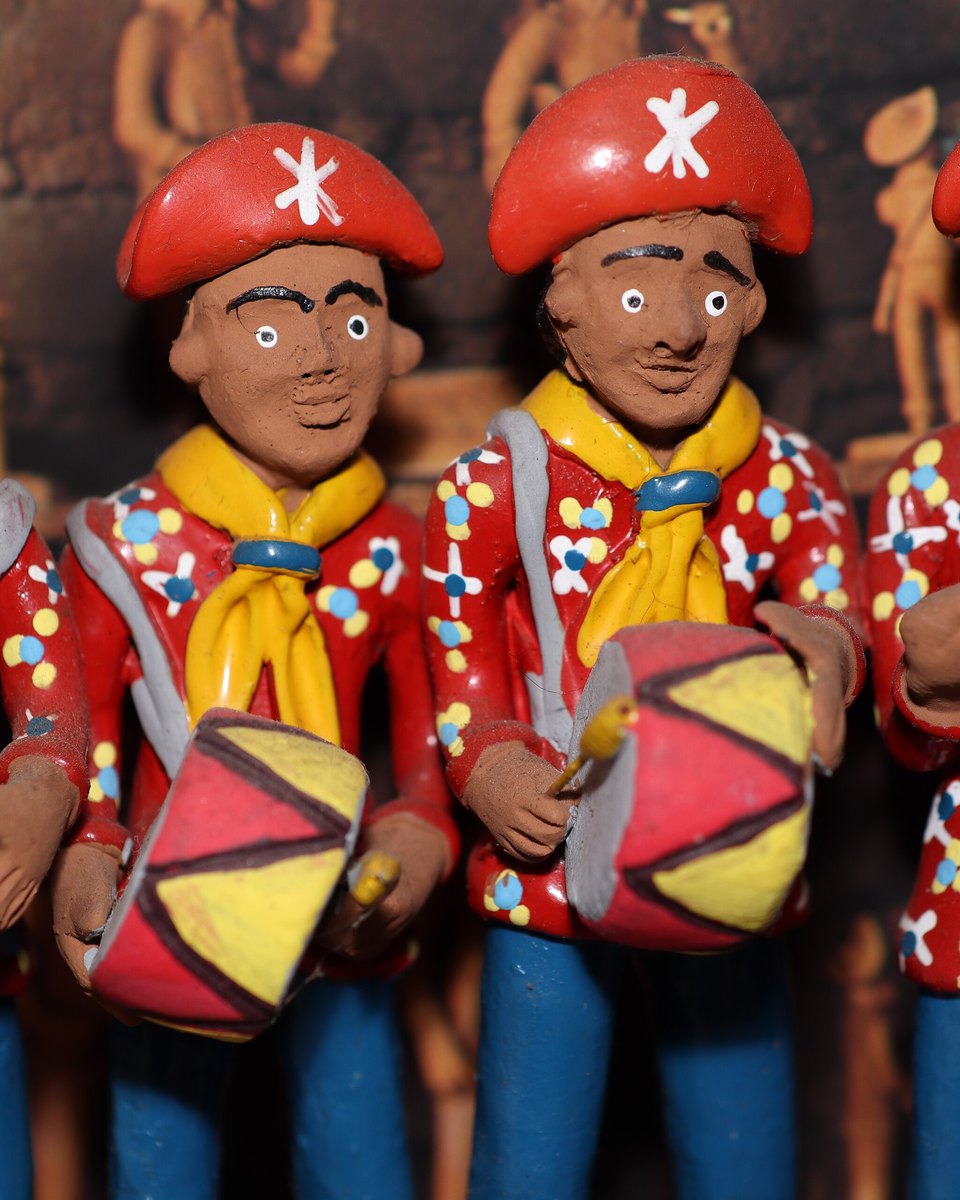Quatro figuras de barro pintadas de tocadores de zabumba e pandeiro, lado a lado, com boina vermelha de estrela branca, lenço amarelo e camisa vermelha de bolinhas.