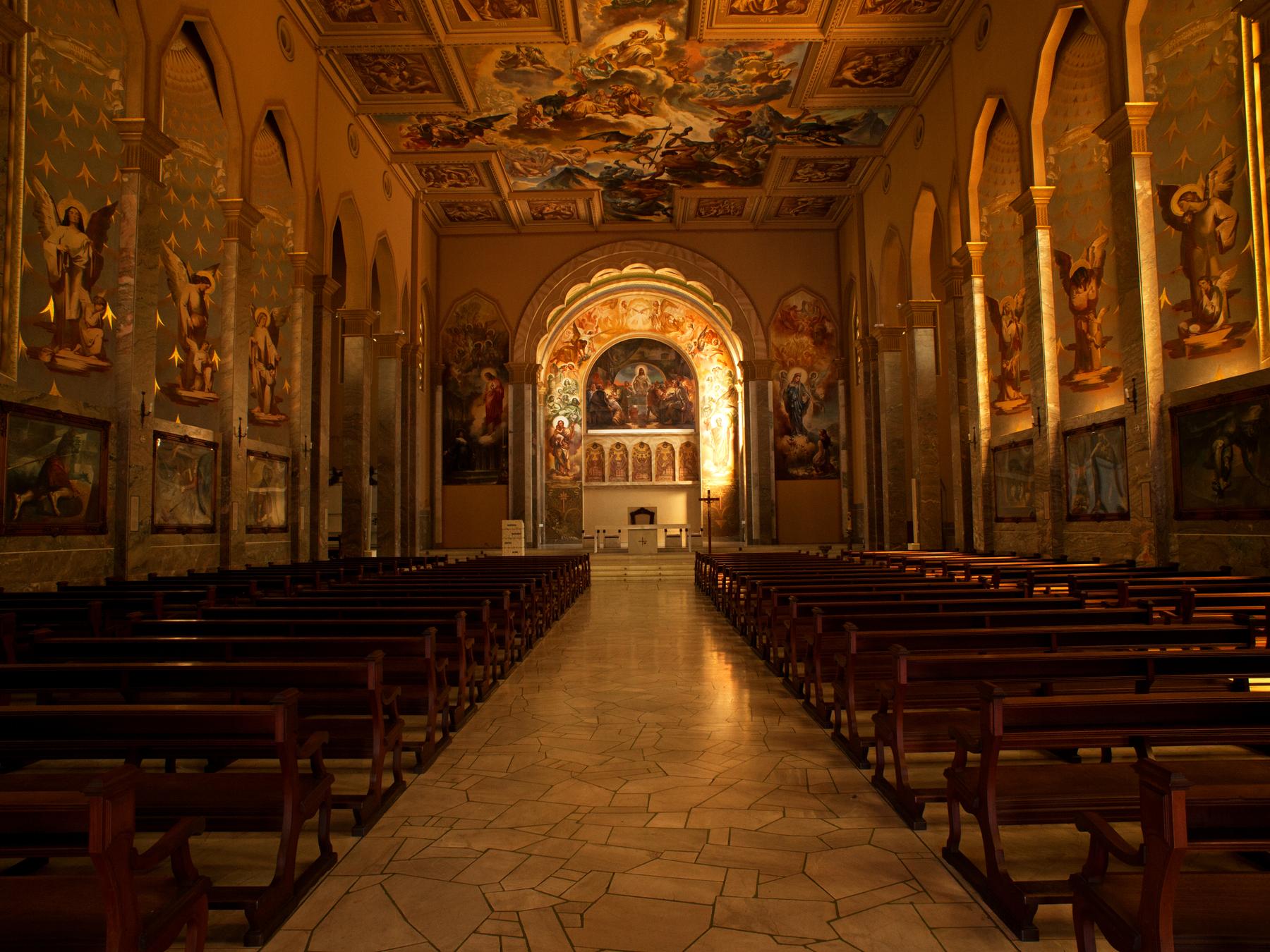 Nave da Igreja de São Pelegrino, em Caxias do Sul, com bancos de madeira dos dois lados do corredor, paredes e teto cobertos de pinturas de Aldo Locatelli e o altar iluminado ao fundo.