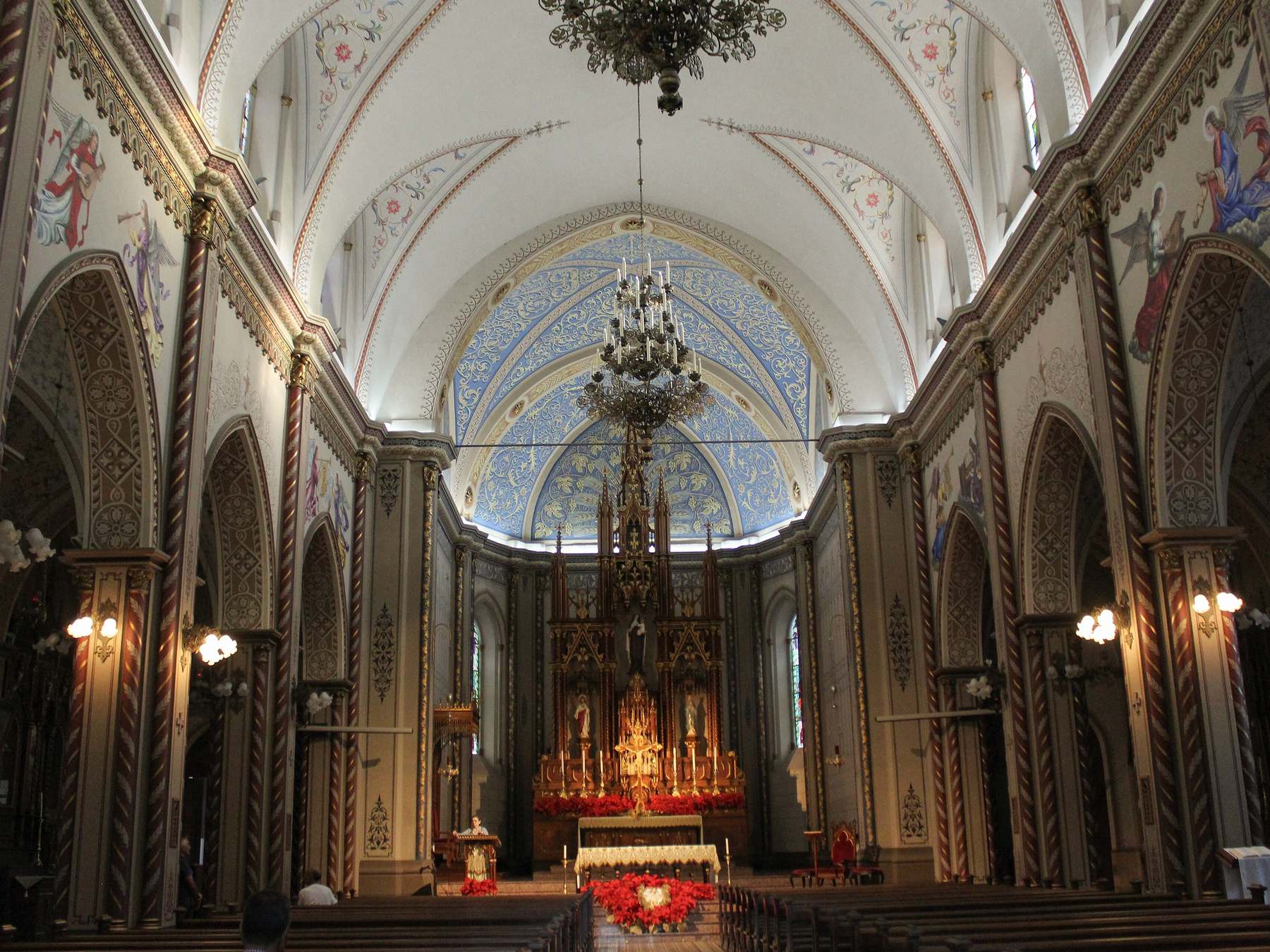 Interior da Catedral de Caxias do Sul, com colunas em arco, lustres acesos, teto pintado de azul com arabescos e o altar de madeira dourada ao fundo.