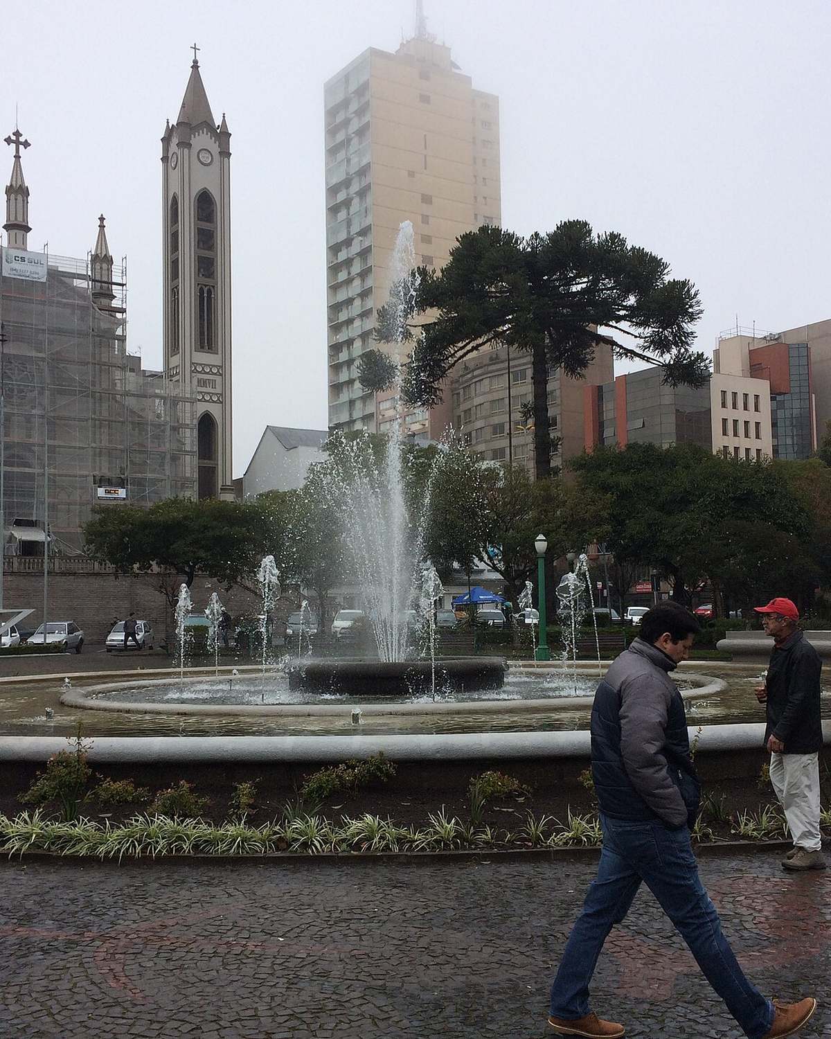 Pedestres de casaco atravessando a Praça Dante Alighieri, em Caxias do Sul, ao lado do chafariz, com a torre da Catedral e prédios do Centro ao fundo, num dia nublado.