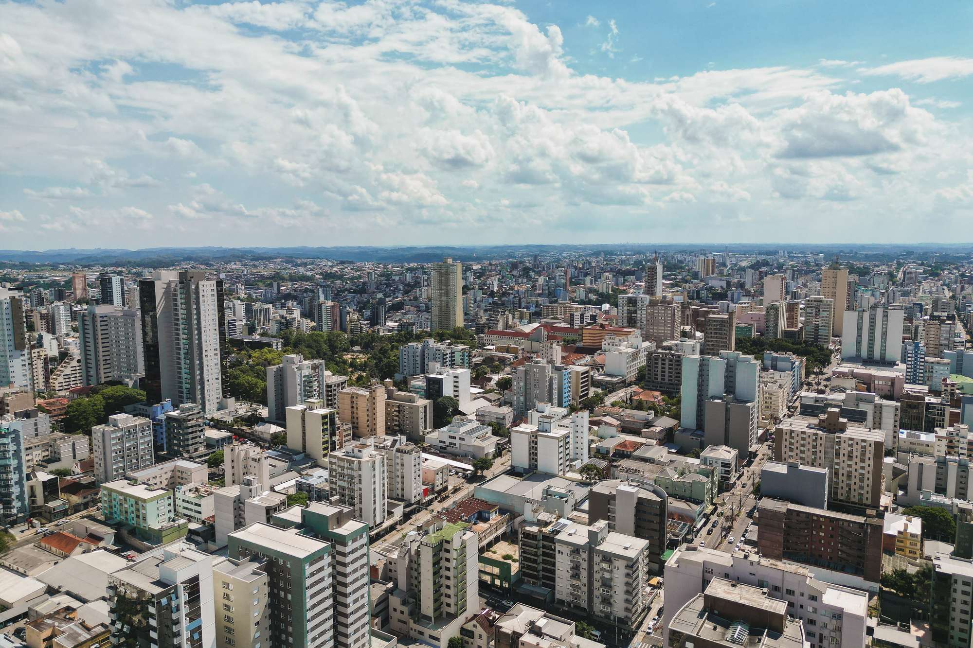 Vista aérea do centro de Caxias do Sul, com prédios altos e ruas em quadrícula até o horizonte, sob céu azul com nuvens brancas.