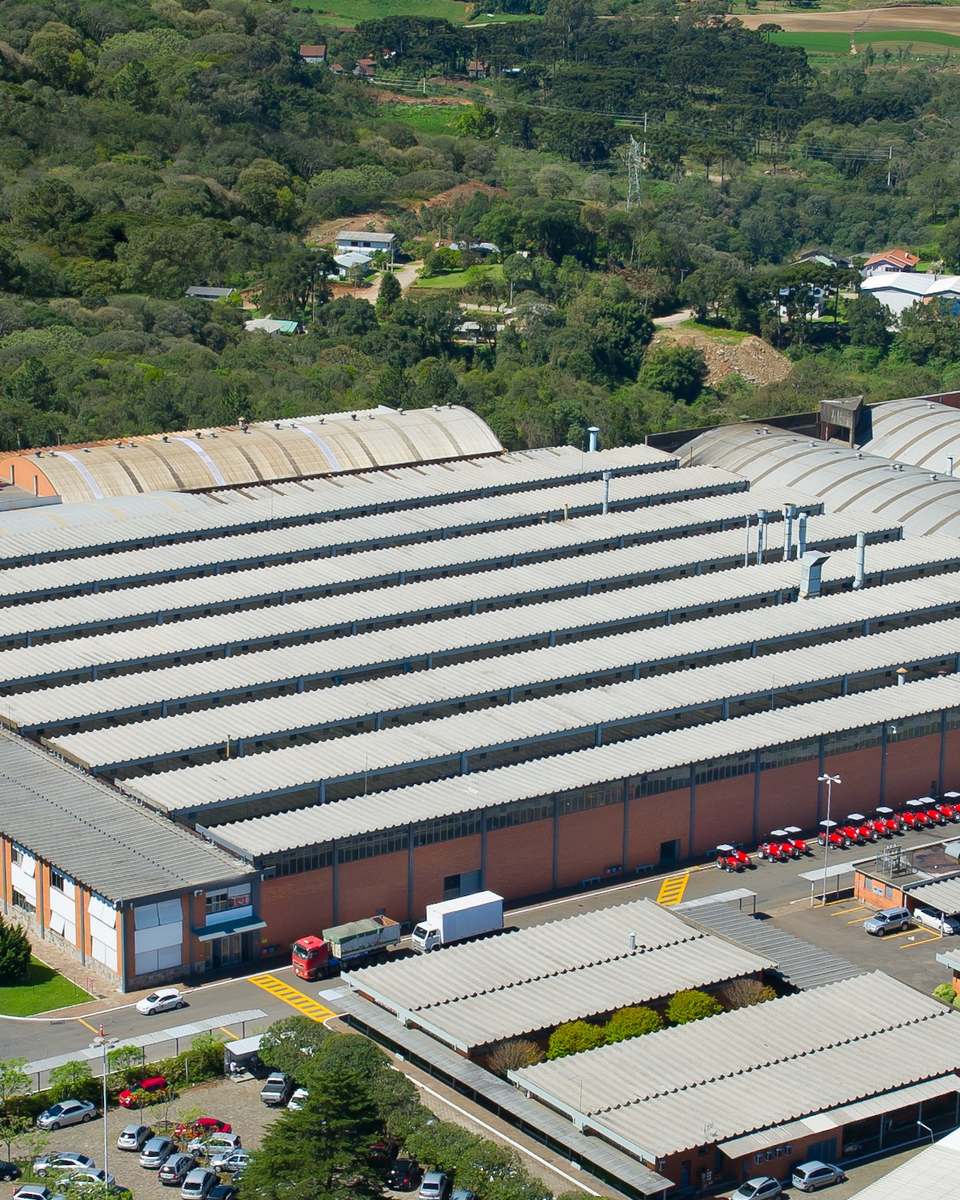 Vista aérea de um grande pavilhão industrial de telhado cinza em Caxias do Sul, com estacionamento, árvores em volta e uma avenida movimentada à esquerda.