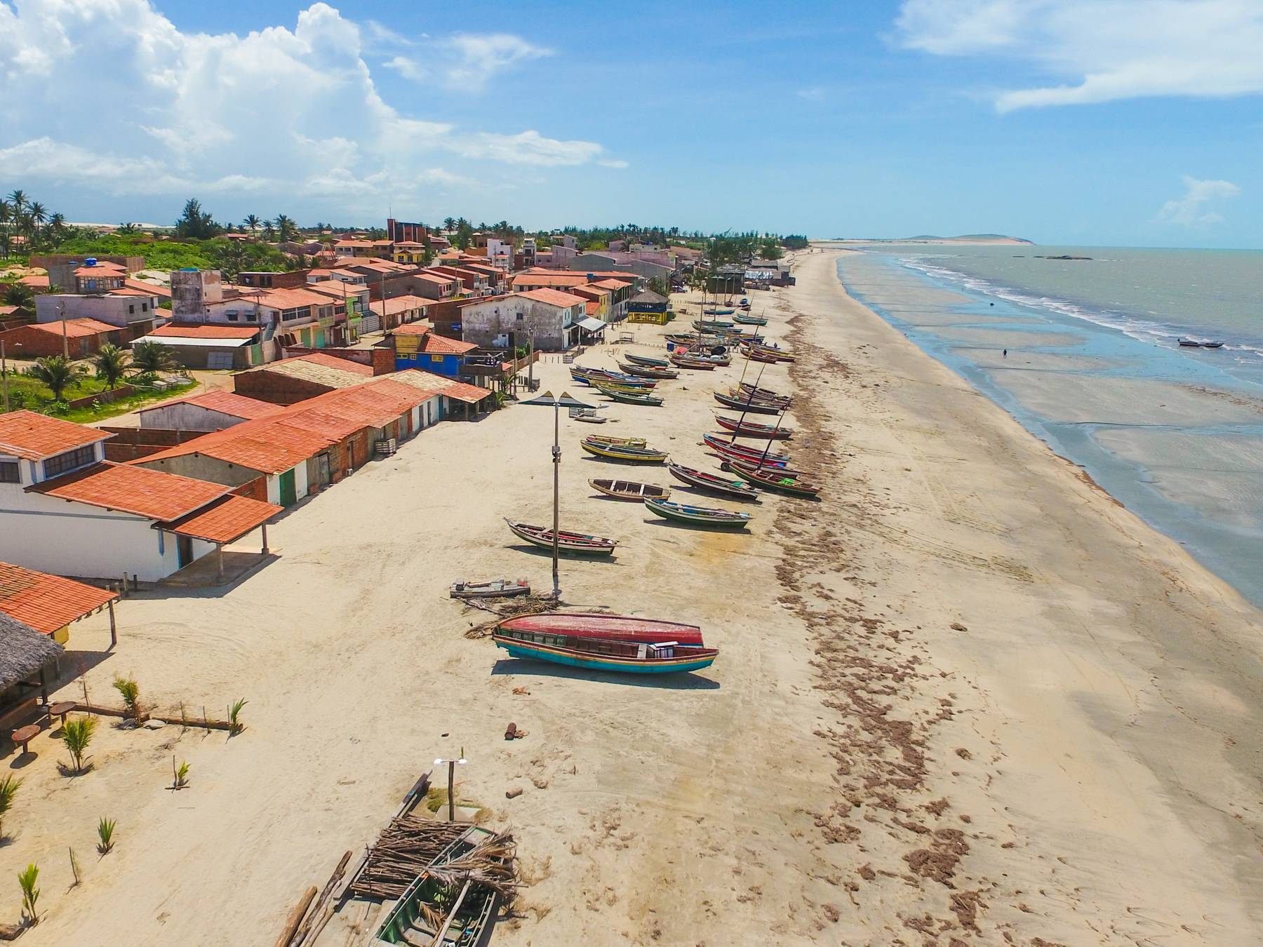 Vista aérea da vila do Preá, com as casas de telhado vermelho de frente para a praia, barcos coloridos na areia e o mar se estendendo em linha reta até o horizonte.