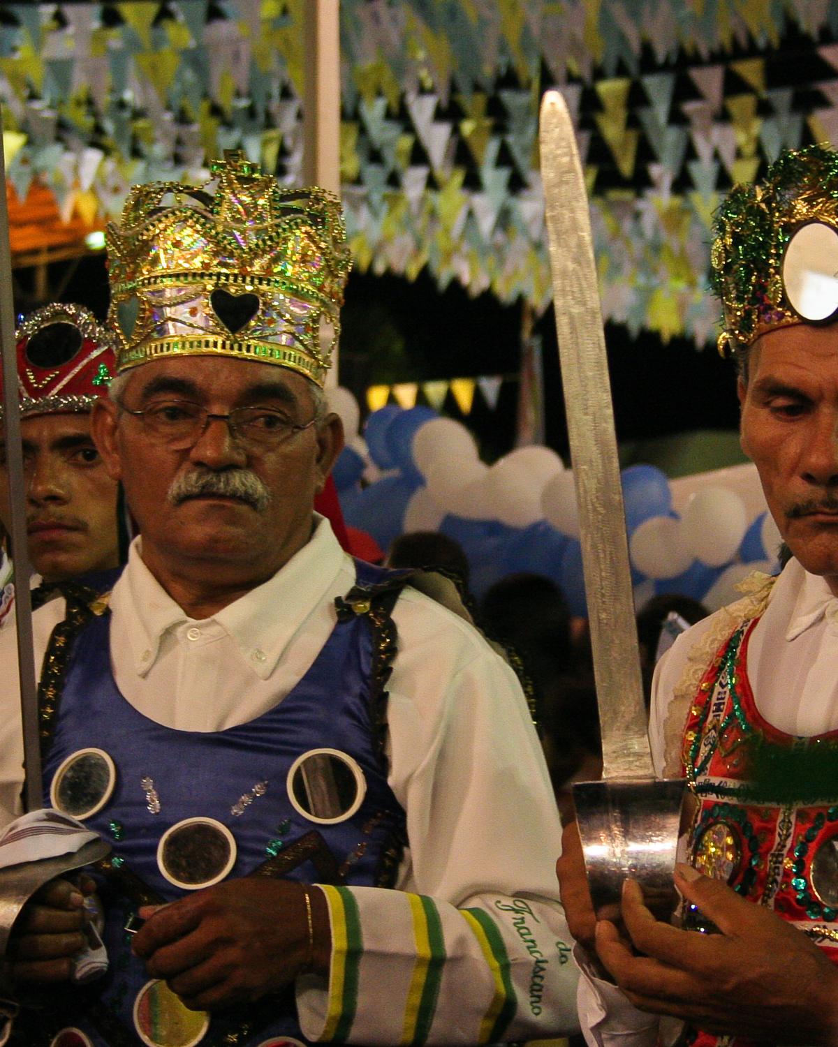Mestres e brincantes de reisado com coroas enfeitadas de espelhos, peitorais bordados e espadas, numa apresentação à noite em Juazeiro do Norte, sob bandeirinhas coloridas.