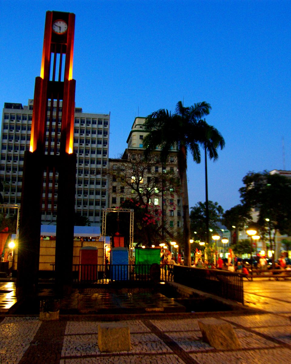 Praça do Ferreira, no Centro de Fortaleza, ao anoitecer, com a Coluna da Hora iluminada, palmeiras, prédios antigos e pessoas caminhando pelo piso de pedra portuguesa.