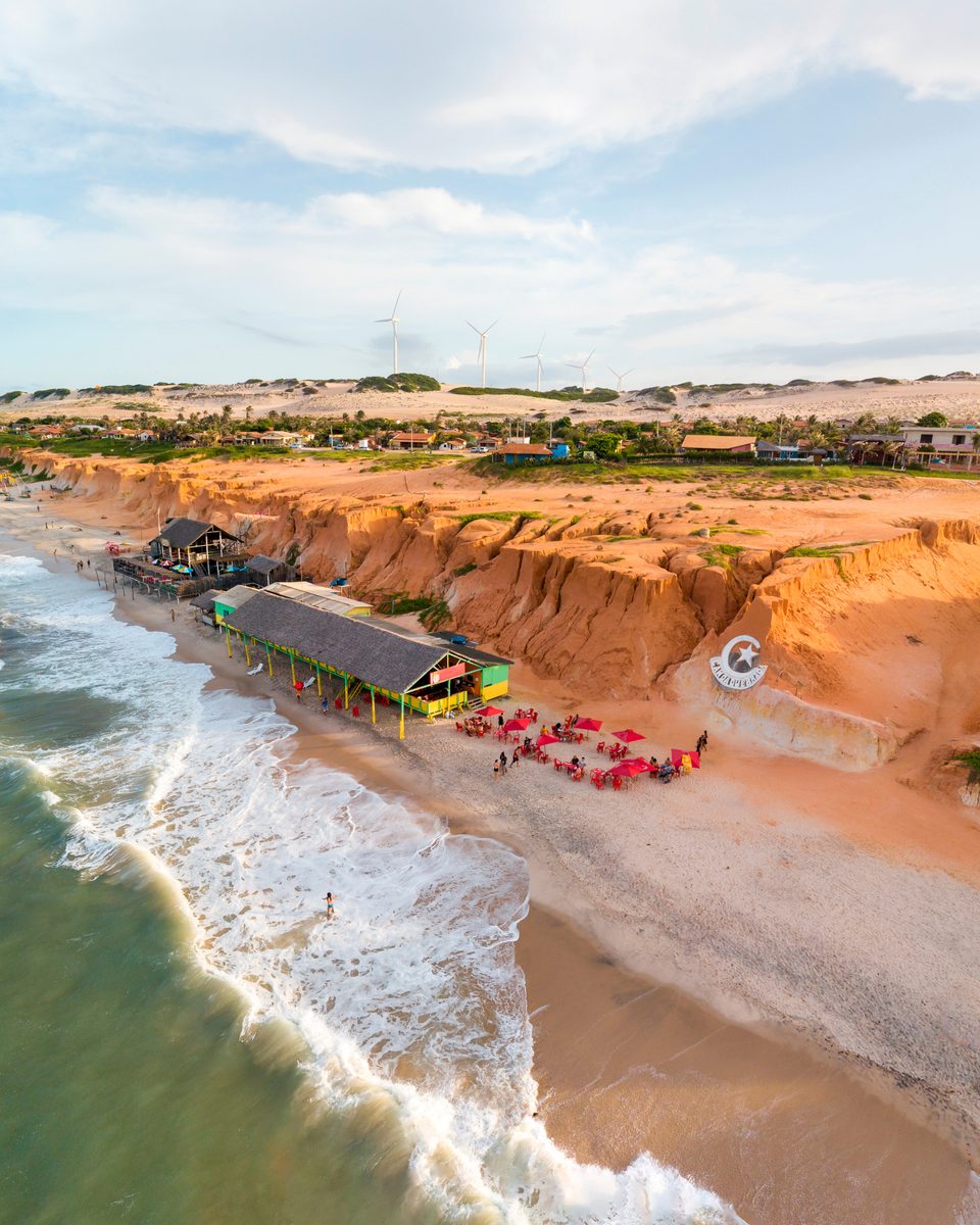 Vista aérea de Canoa Quebrada, em Aracati, com as falésias avermelhadas, as barracas de praia na areia, as ondas quebrando e as dunas ao fundo.