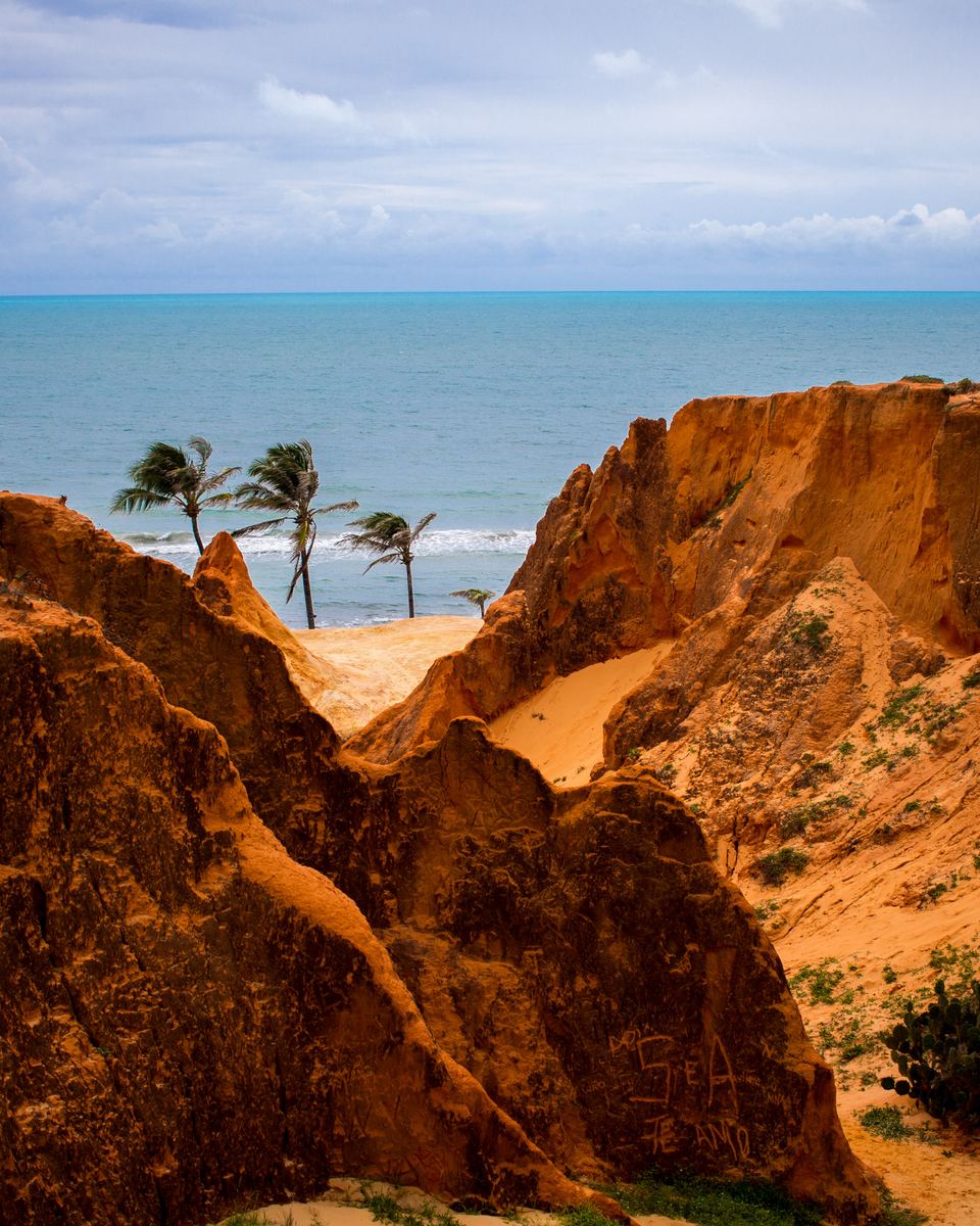 Falésias de areia avermelhada e alaranjada do Morro Branco, em Beberibe, com coqueiros no alto e o mar azul ao fundo sob céu com nuvens.