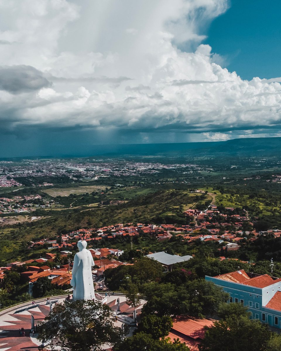 Estátua branca do padre Cícero no alto da Colina do Horto, em Juazeiro do Norte, vista de drone, com a cidade de Juazeiro espalhada pela planície ao fundo e nuvens de chuva no céu.