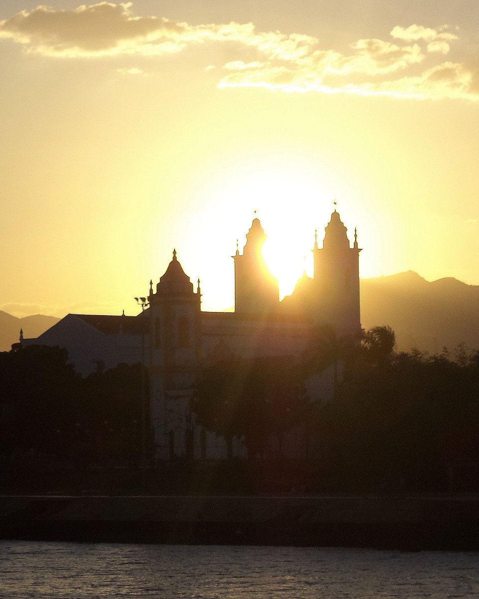 Silhueta da catedral de Sobral e do casario à beira do Rio Acaraú ao pôr do sol, com o céu dourado e a serra ao fundo.
