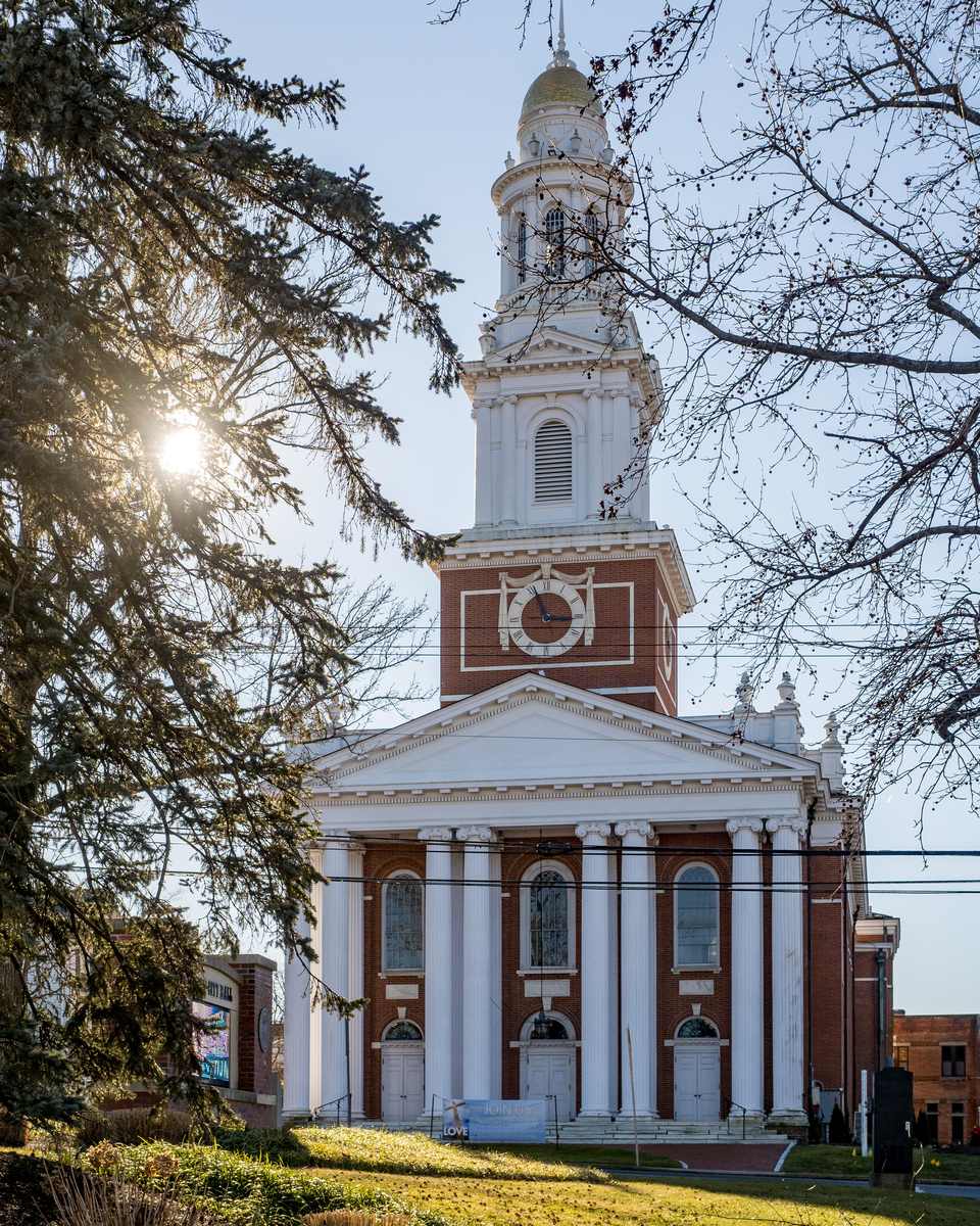 Igreja de tijolos com colunas brancas e torre branca em Danbury, com o sol atravessando os galhos das árvores.