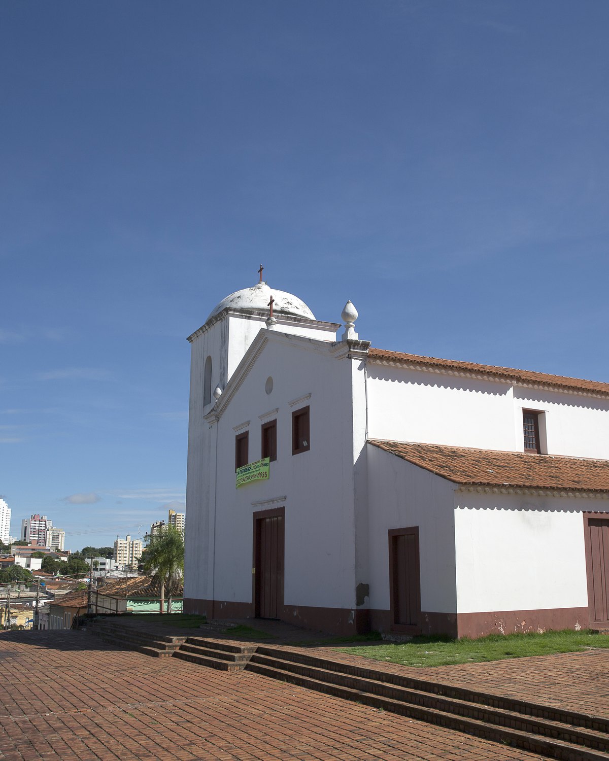Igreja de Nossa Senhora do Rosário e São Benedito, branca e de telhado de barro, no alto do largo de piso vermelho, com prédios de Cuiabá ao fundo e céu azul.