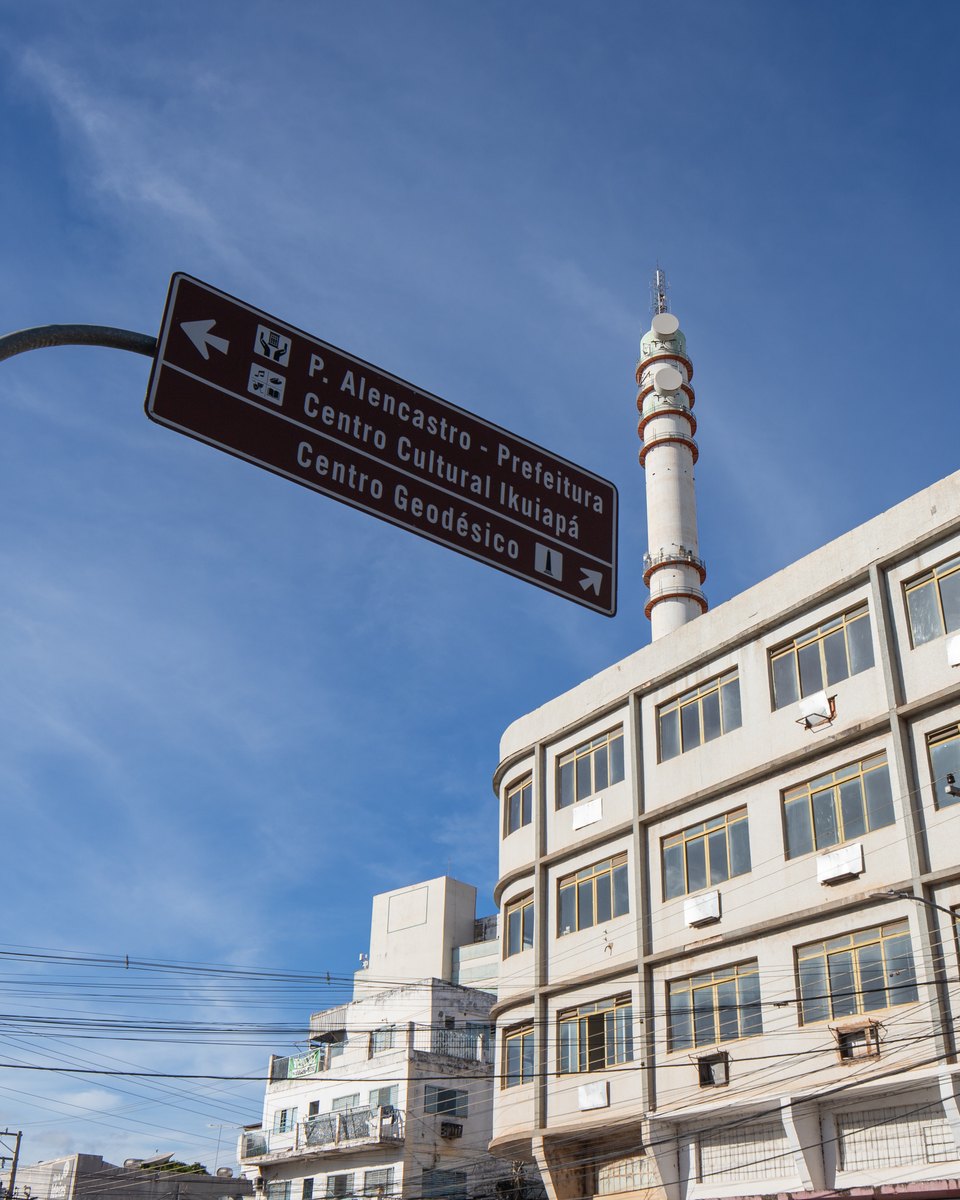 Placa marrom de sinalização turística apontando para a Prefeitura e para o Centro Geodésico, numa esquina de Cuiabá, sob sol forte e céu azul.