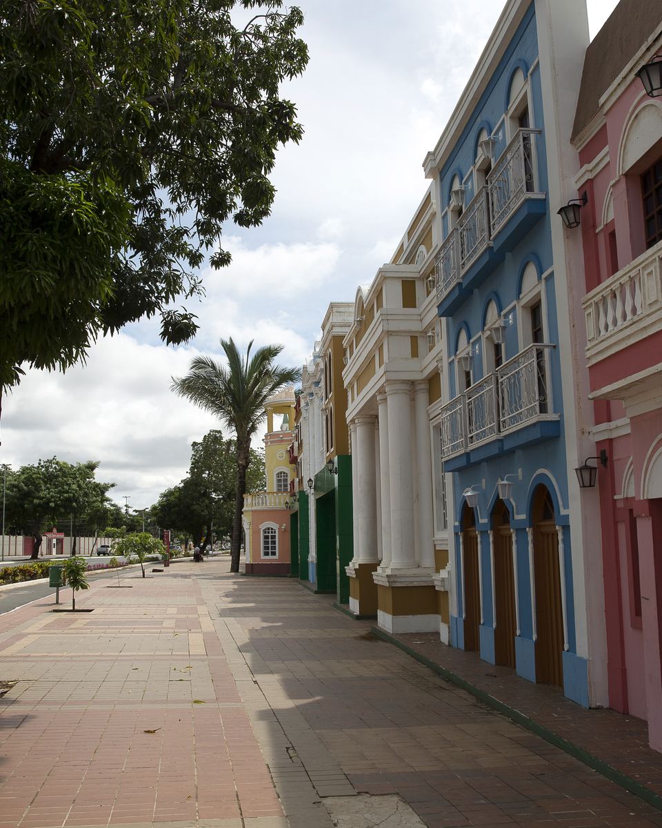 Calçadão arborizado da Orla do Porto, em Cuiabá, com casario colorido de sacada à direita e uma árvore grande à esquerda.