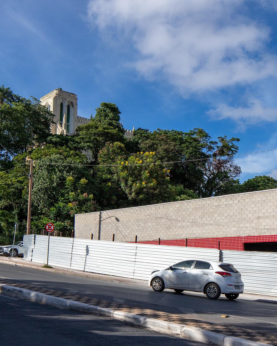 Rua no pé do Morro do Seminário, em Cuiabá, com pedestres na calçada, carros passando e a torre da igreja entre as árvores no alto.