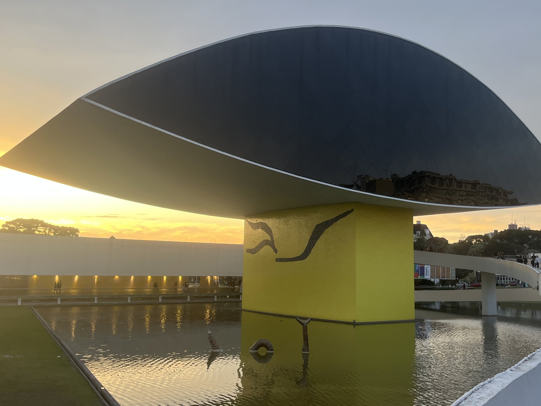 Museu Oscar Niemeyer ao pôr do sol, com a grande estrutura em forma de olho sobre o pilar amarelo pintado e o espelho d'água refletindo o céu dourado.