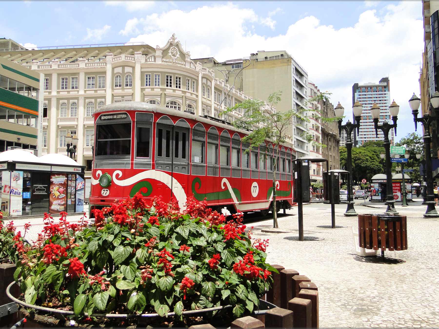 Bondinho vermelho com desenhos brancos e verdes estacionado no calçadão da Rua XV de Novembro, com canteiro de flores vermelhas na frente, postes antigos e um casarão claro ao fundo num dia de sol.