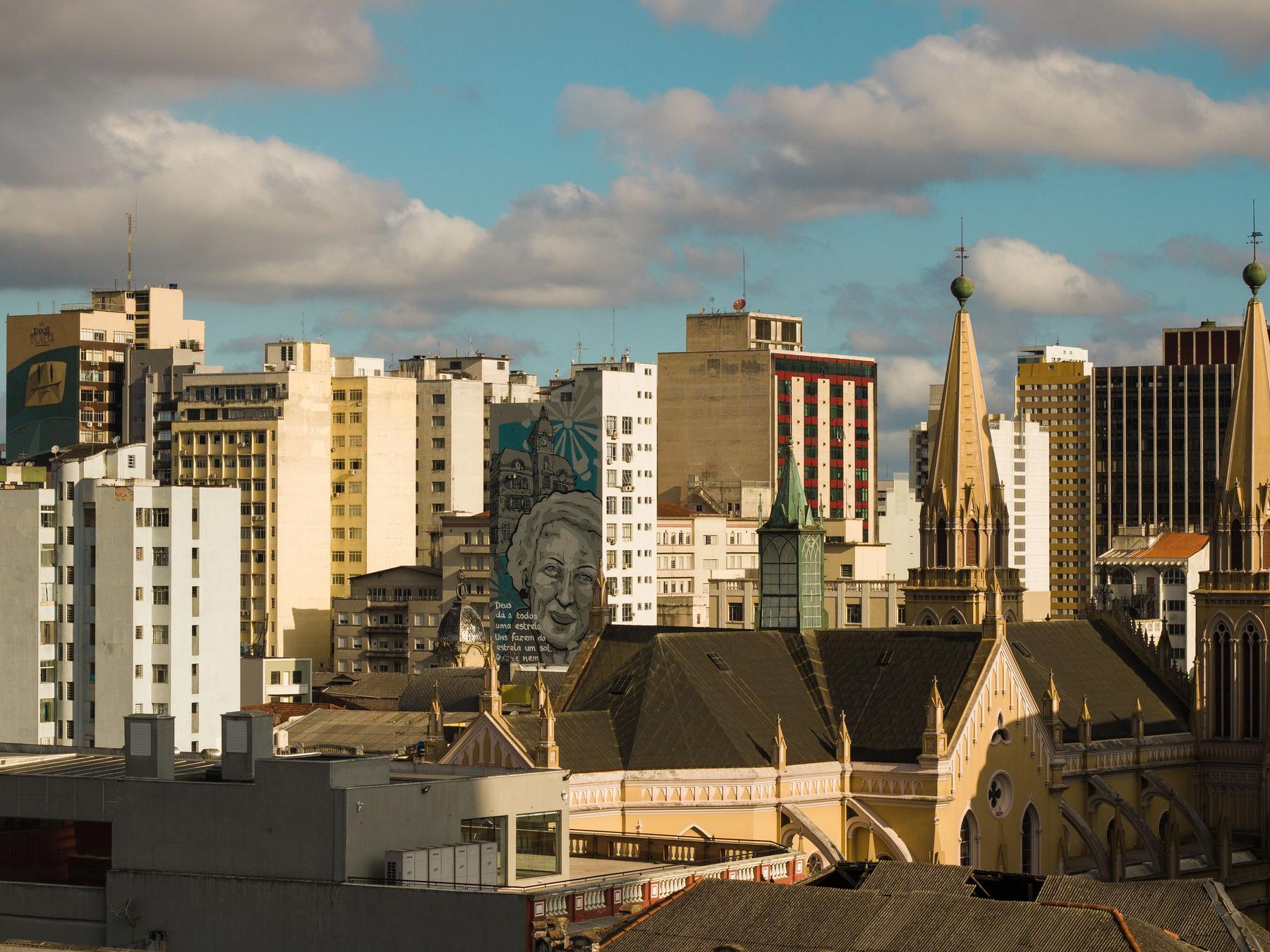 Torres góticas da Catedral de Curitiba iluminadas pelo sol baixo, entre prédios do centro e um grande mural de rosto de mulher, sob céu azul com nuvens.
