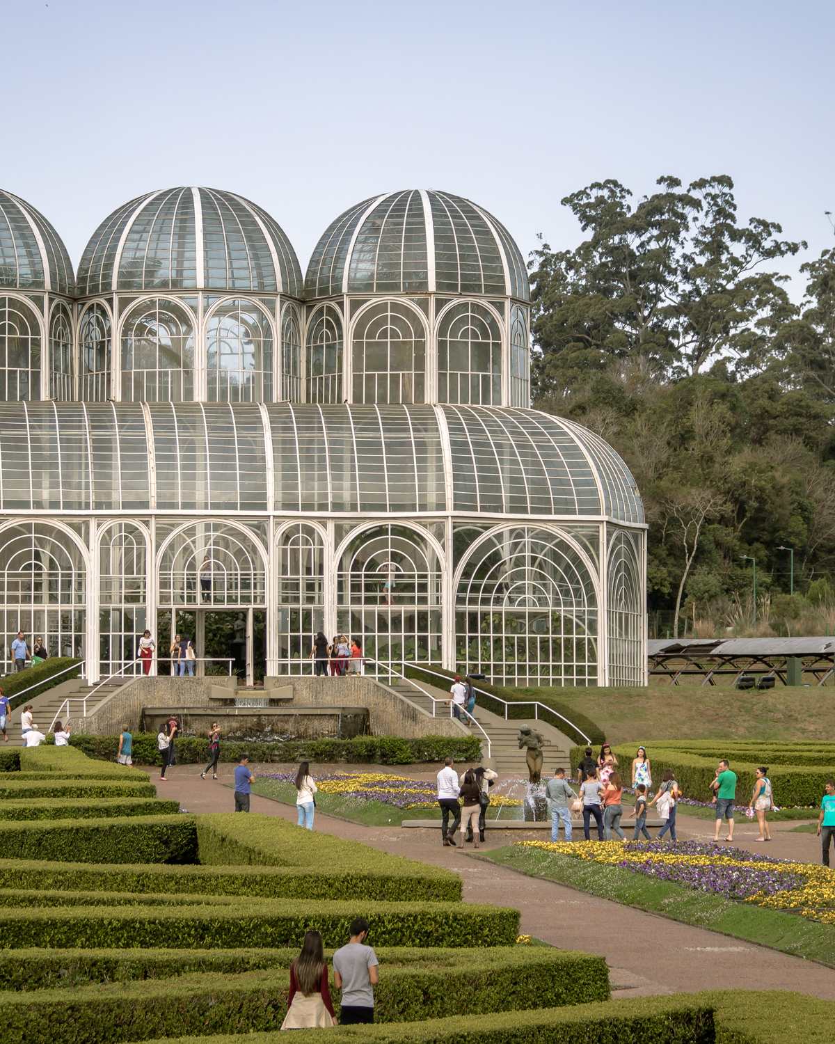 Visitantes caminhando entre os canteiros geométricos do Jardim Botânico de Curitiba, com a estufa de vidro e ferro ao fundo, num dia de sol.