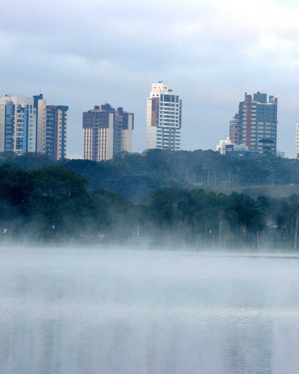 Névoa espessa sobre o lago do Parque Barigui numa manhã fria, com a margem arborizada e os prédios de Curitiba aparecendo acima da neblina.