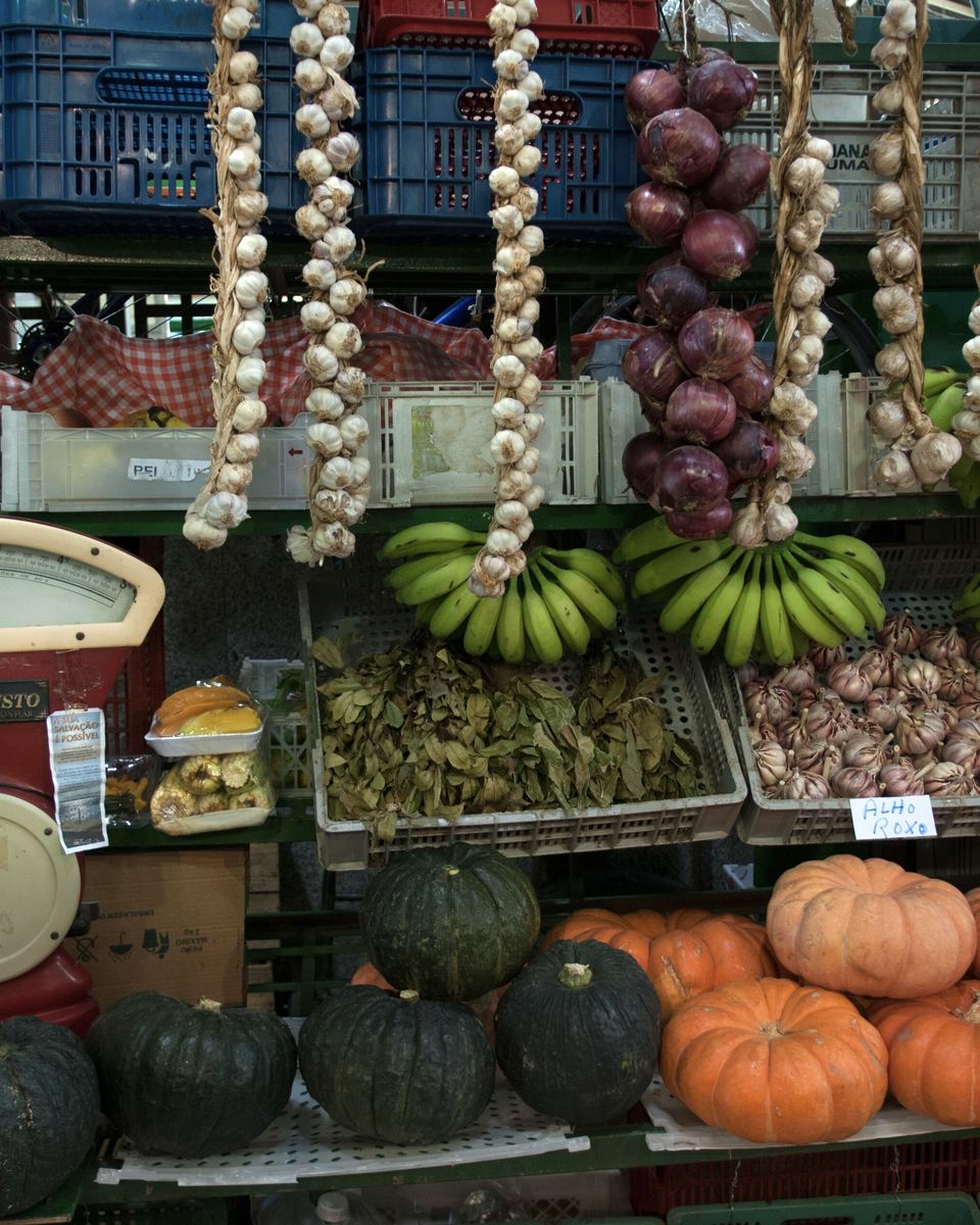 Banca do Mercado Municipal de Curitiba com réstias de alho e cebola penduradas, abóboras, bananas e outros legumes arrumados no balcão.