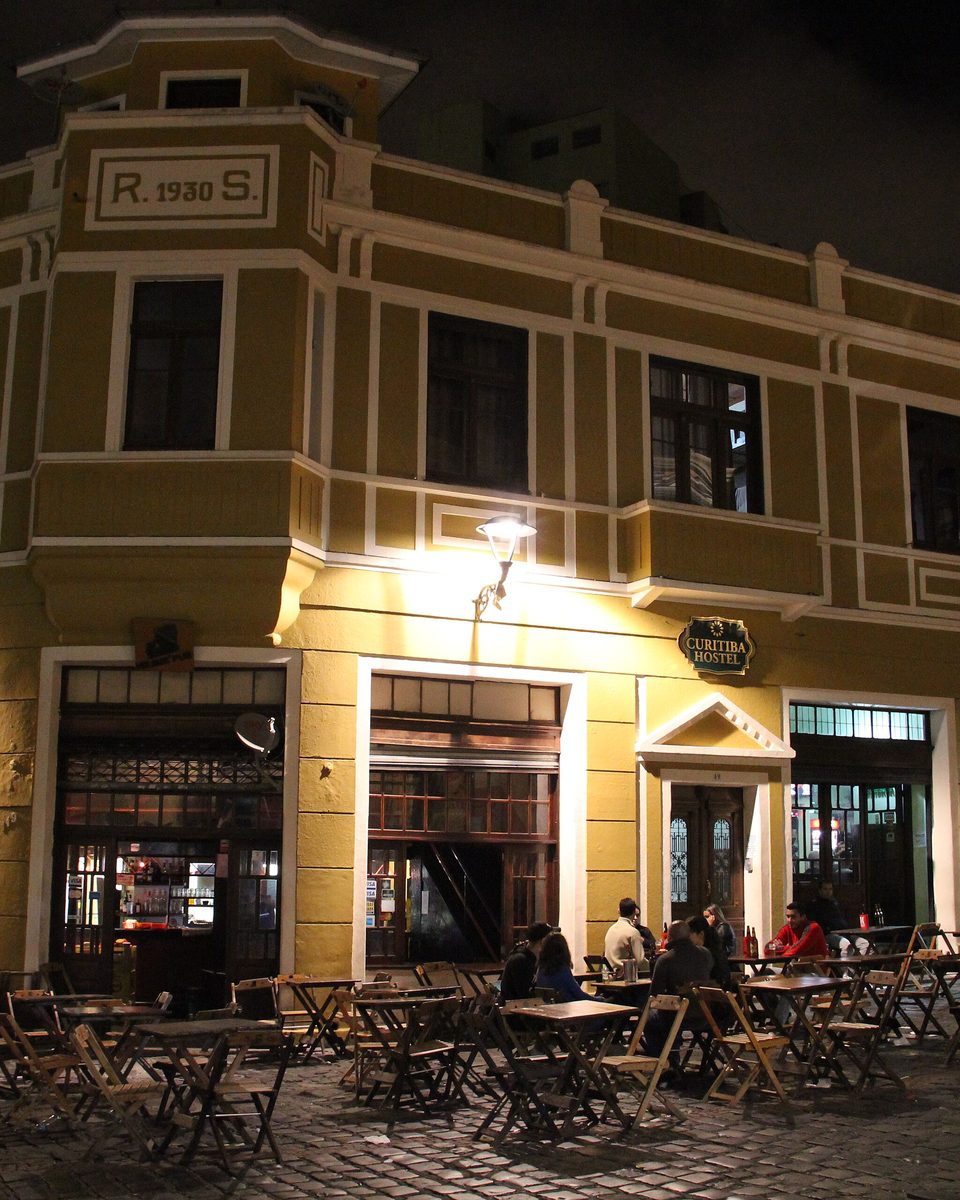 Pessoas nas mesas da calçada de um bar instalado num casarão amarelo do Largo da Ordem, em Curitiba, à noite, com o calçamento de pedra iluminado.