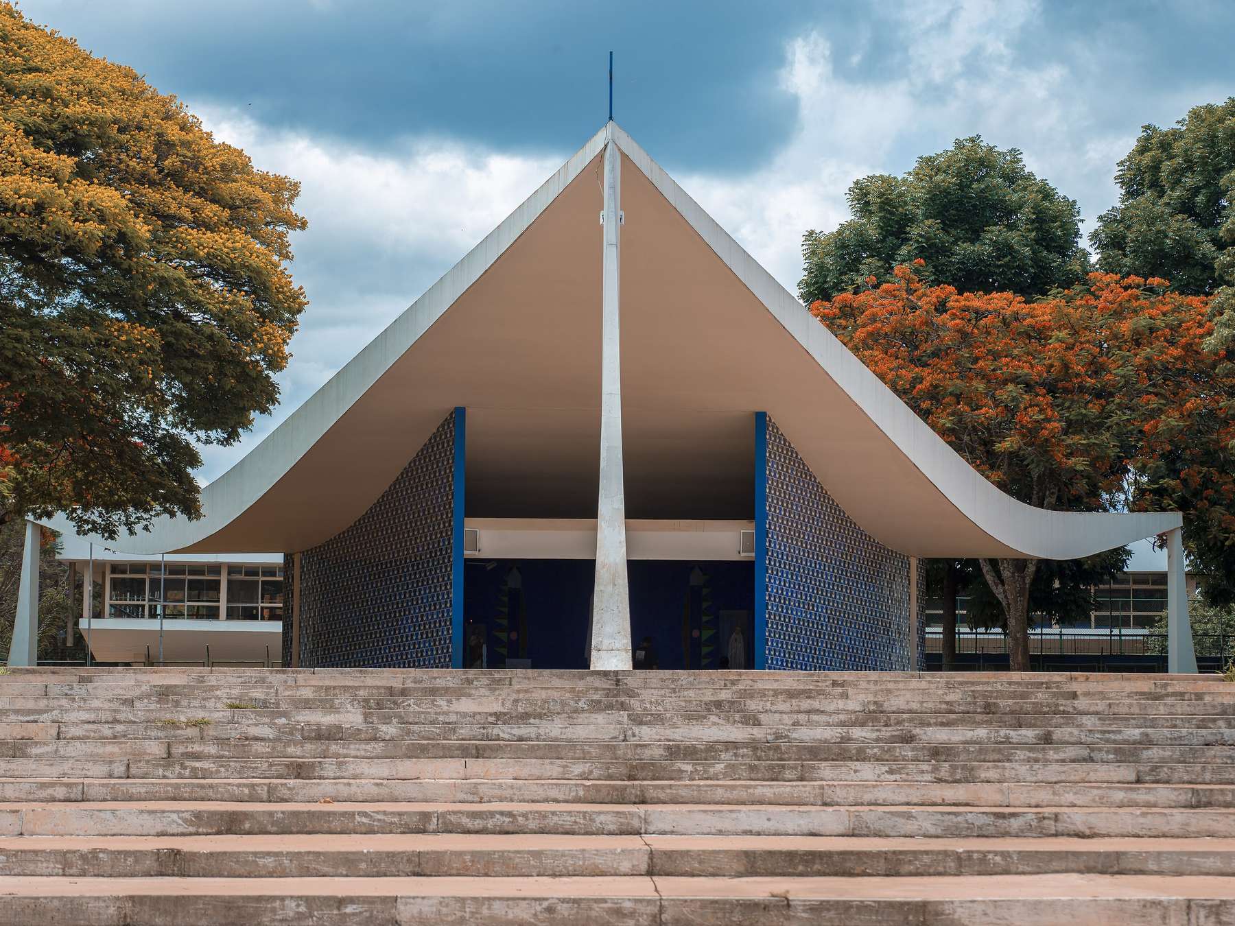 Igrejinha Nossa Senhora de Fátima, de telhado branco em forma de chapéu apoiado em colunas, com escadaria de concreto à frente e árvores floridas em volta.