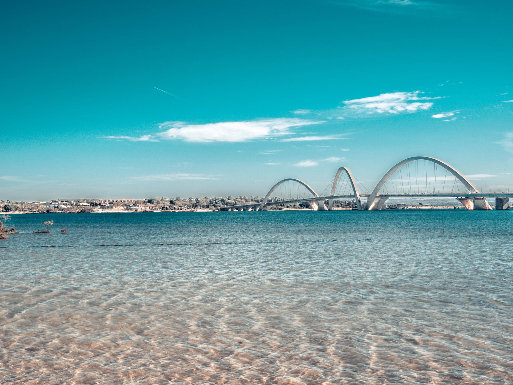 Orla do Lago Paranoá com areia clara e água azul-turquesa em primeiro plano e a Ponte JK atravessando o lago ao fundo, sob céu azul.
