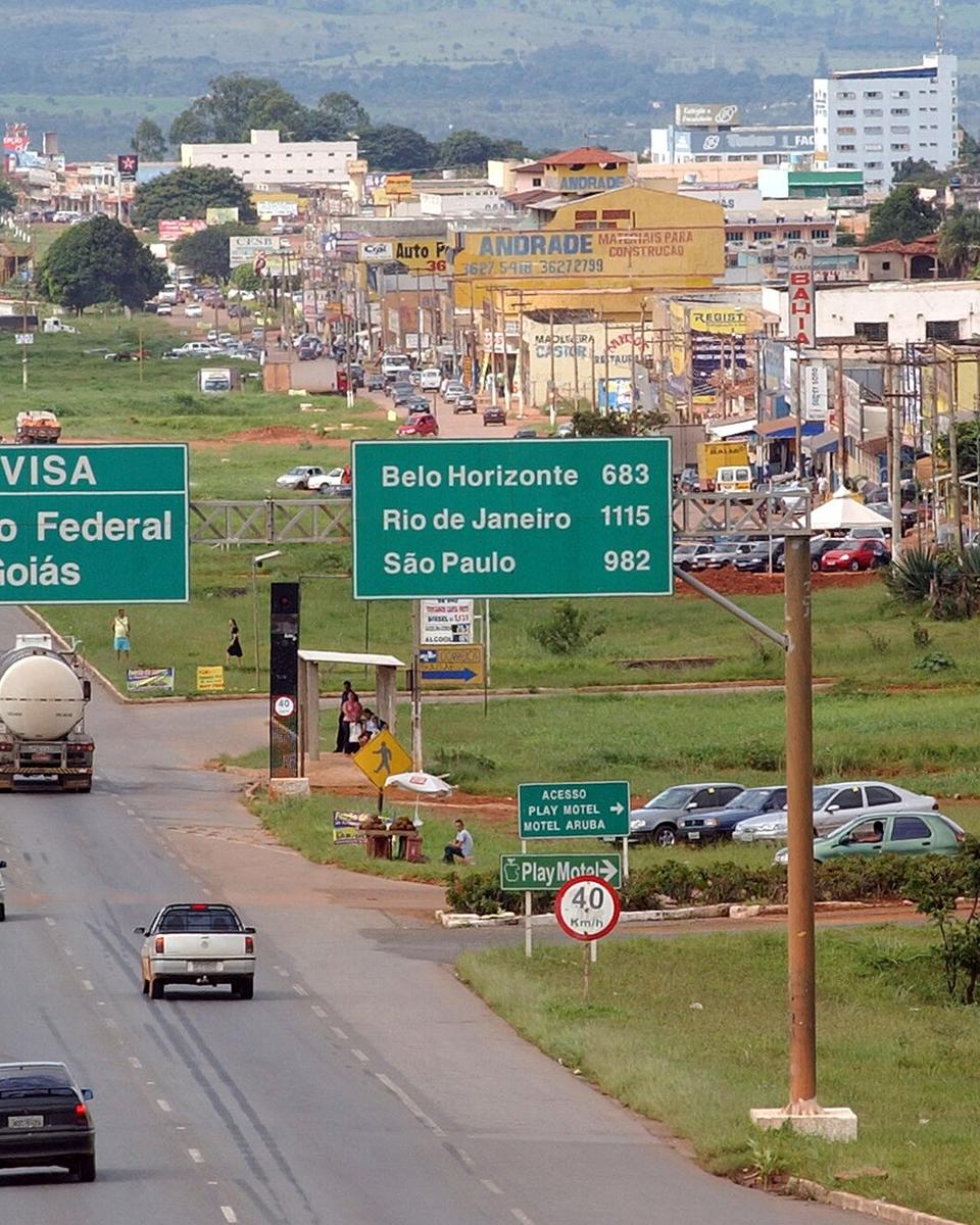 Rodovia com placa verde de divisa entre o Distrito Federal e Goiás, caminhão-tanque e carros na pista, e casas e comércio de uma cidade do Entorno ao fundo.