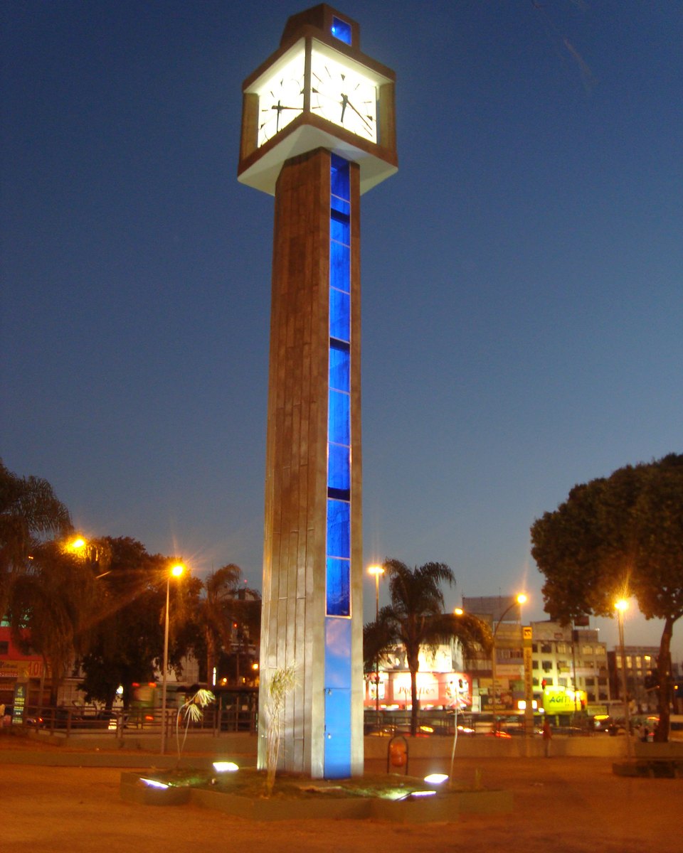 Torre do Relógio da Praça Central de Taguatinga iluminada ao anoitecer, com o mostrador aceso no alto, palmeiras e as luzes da cidade em volta.