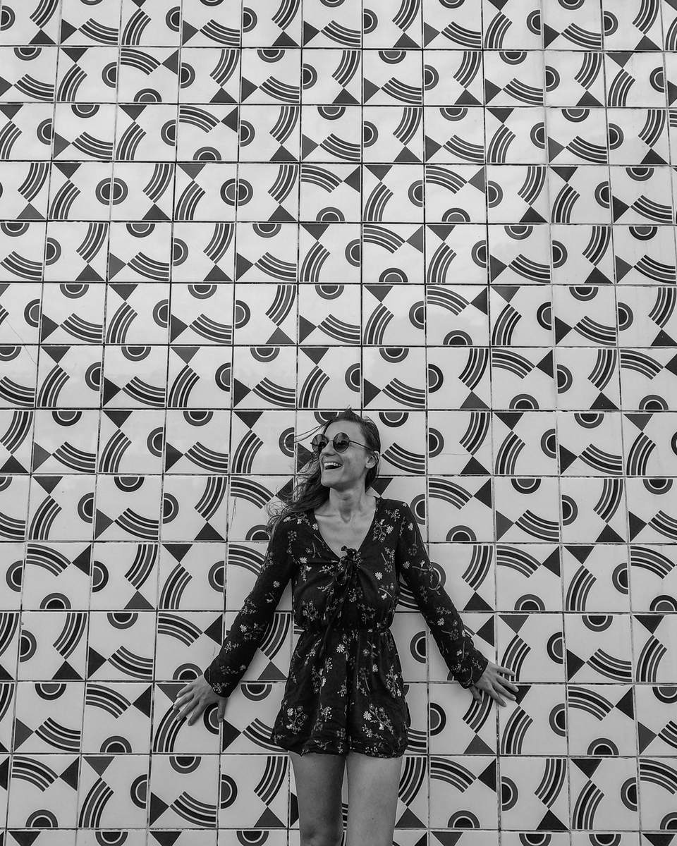 Mulher de vestido estampado em pé, de braços abertos, diante de um grande painel de azulejos geométricos em preto e branco de Athos Bulcão, na Universidade de Brasília.