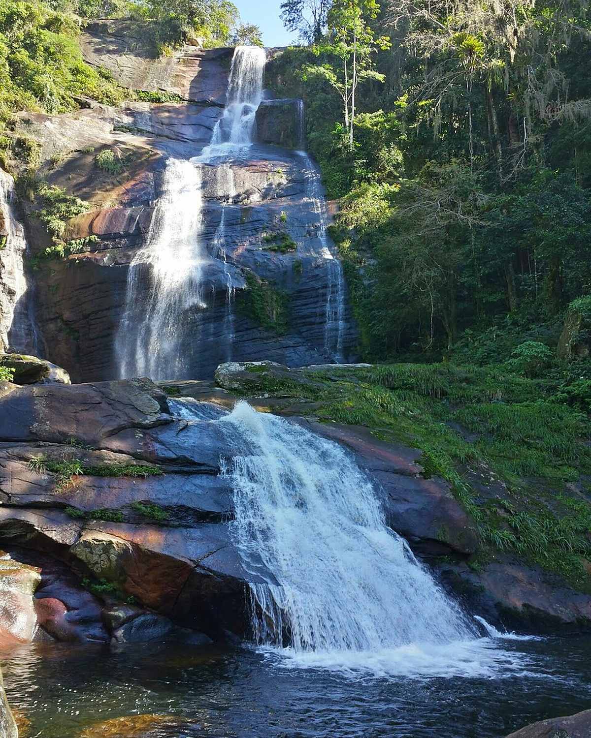 Cachoeira em dois saltos descendo por pedras entre a mata atlântica, formando um poço de água escura em Xerém.