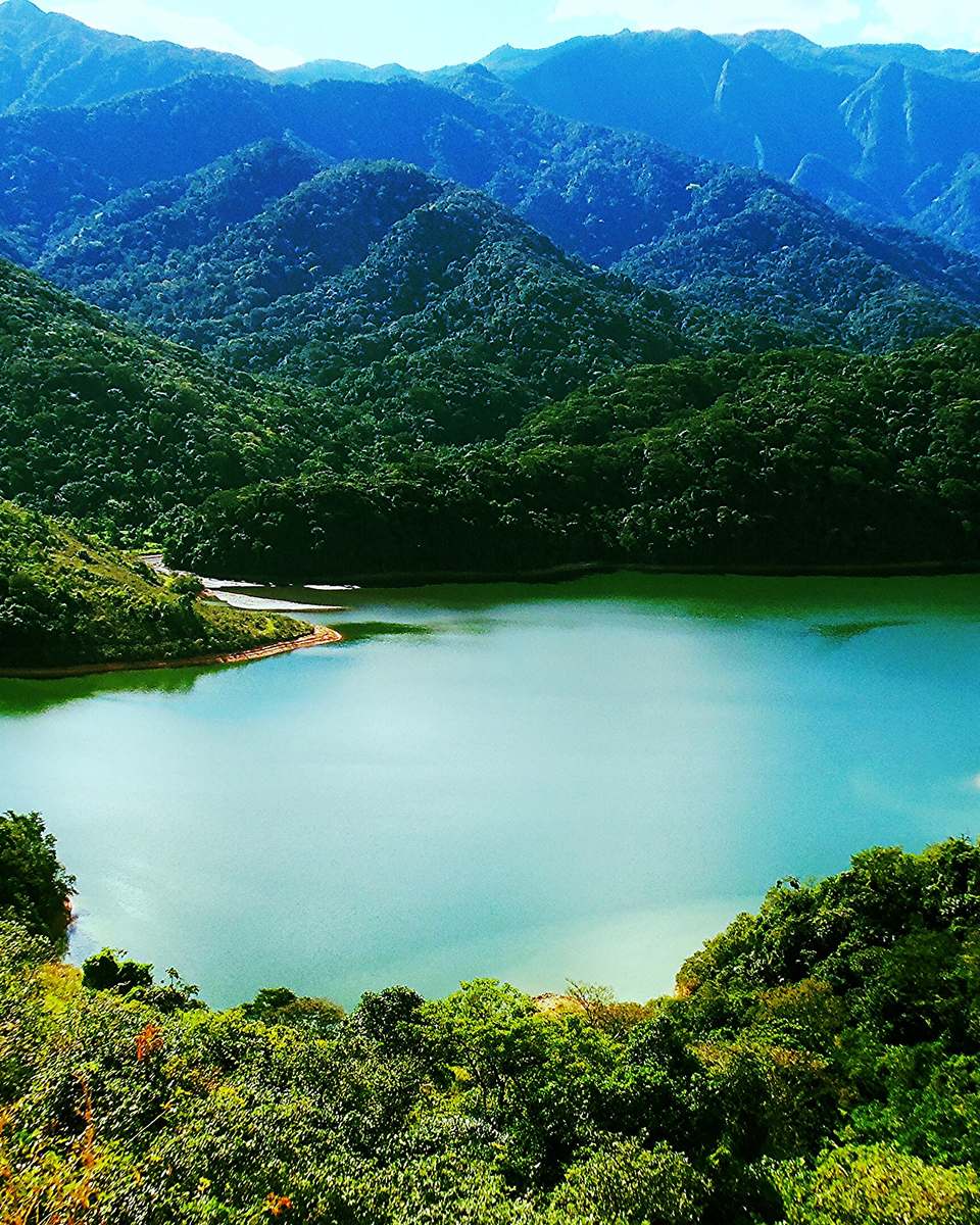 Represa do Garrão, em Xerém, com a água verde-azulada cercada de morros cobertos de mata atlântica e as montanhas da serra ao fundo, sob céu azul.