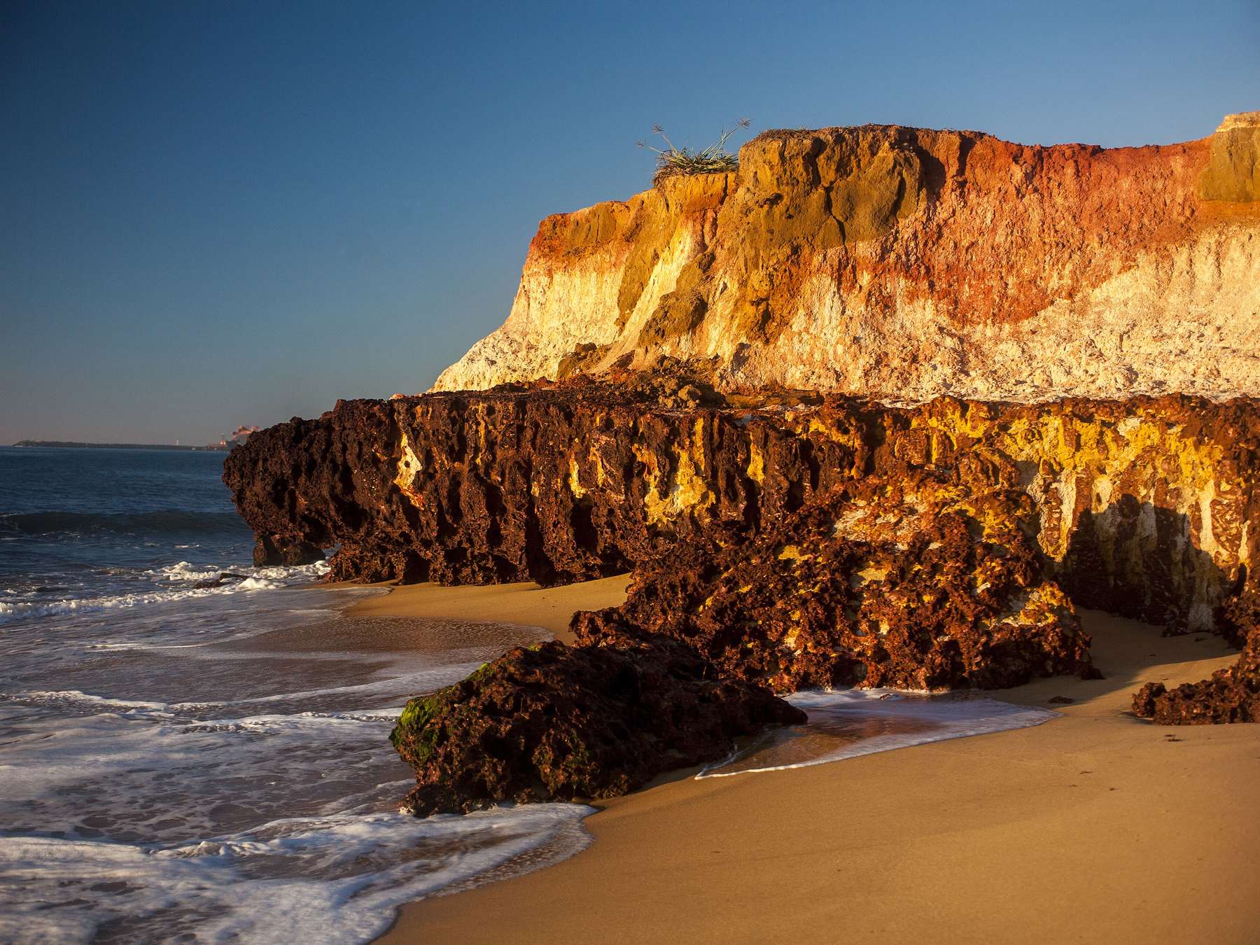 Falésias de areia dourada e avermelhada à beira-mar em Anchieta, iluminadas pelo sol baixo, com a faixa de areia e as ondas quebrando no primeiro plano.