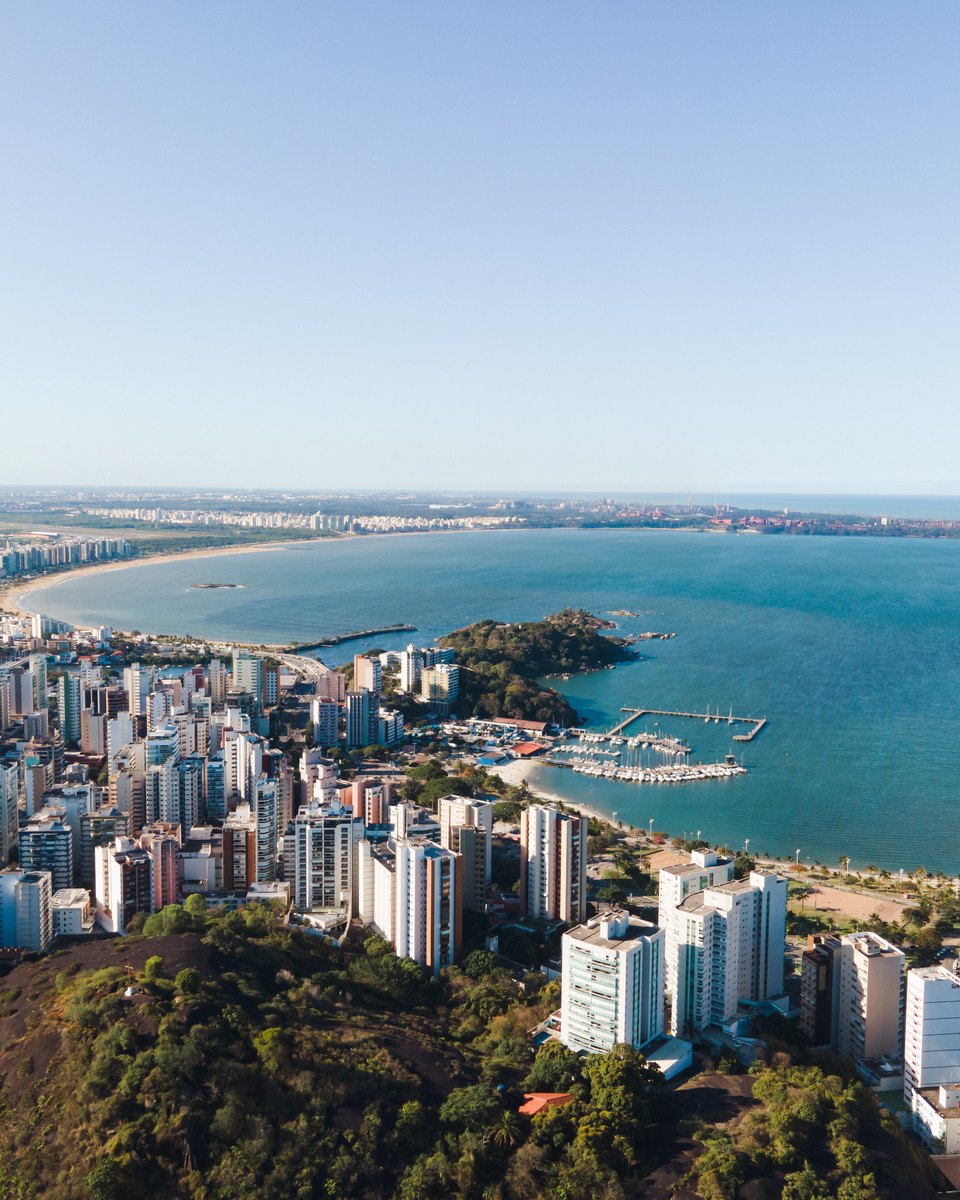 Vista aérea de Vitória com prédios, marina, baía e a praia de Camburi ao fundo.